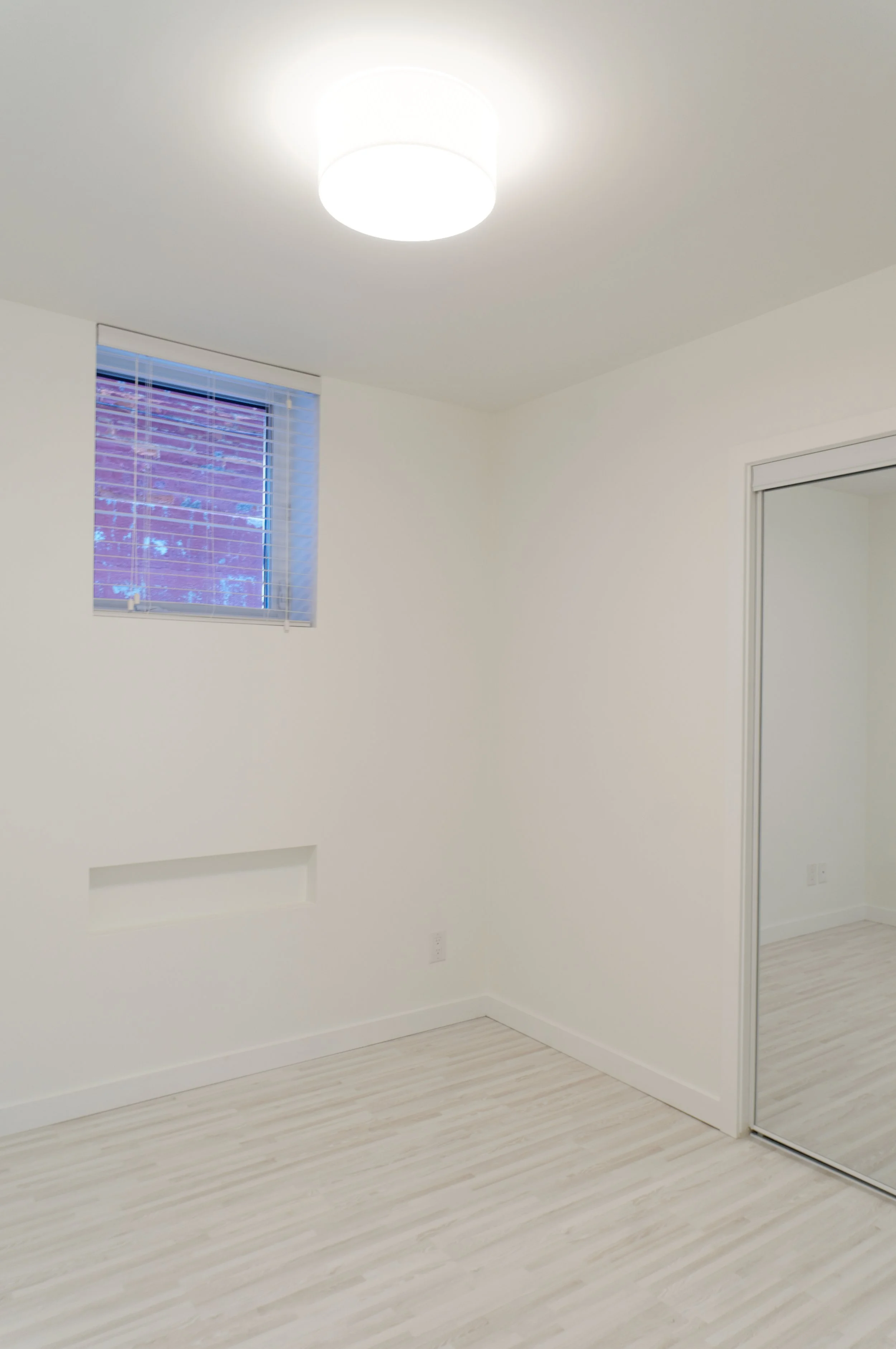 Empty room with white walls, a window with blinds, a ceiling light, and a sliding mirror closet door.