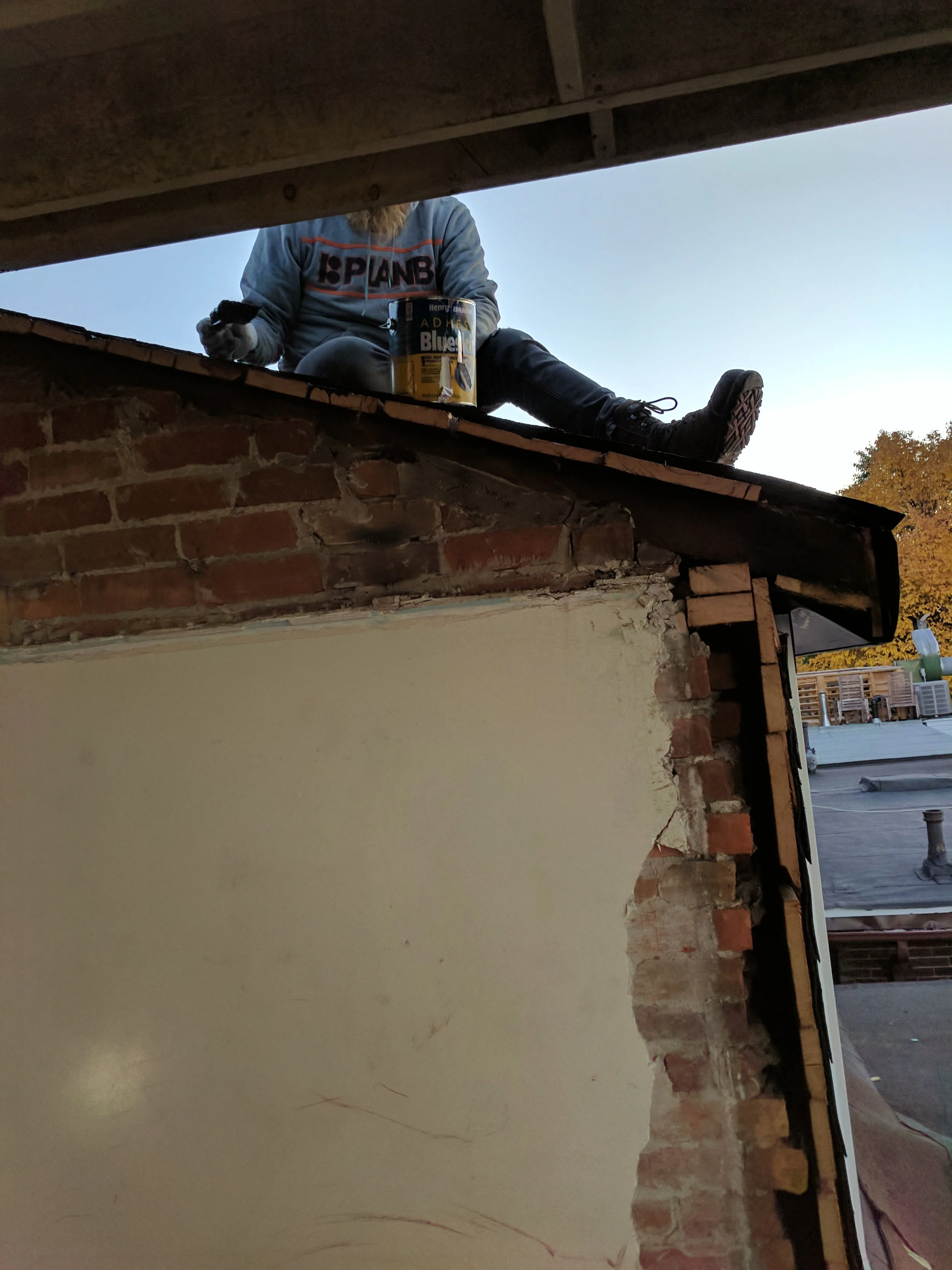 A person working on a roof, seated on the edge, with a can of Henry Adams Blue paint nearby. The photo is taken from below, showing the person’s legs and part of the upper body, with a clear sky and some autumn trees in the background.