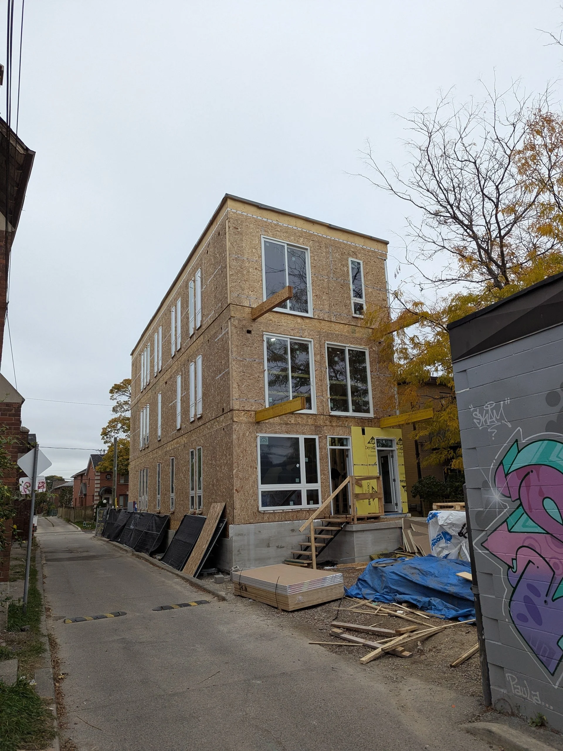 A multi-story building under construction with plywood exterior walls and large windows. Construction materials and equipment are seen in the foreground, including stacks of plywood, blue tarps, and scattered wood. A graffiti-covered wall is on the r