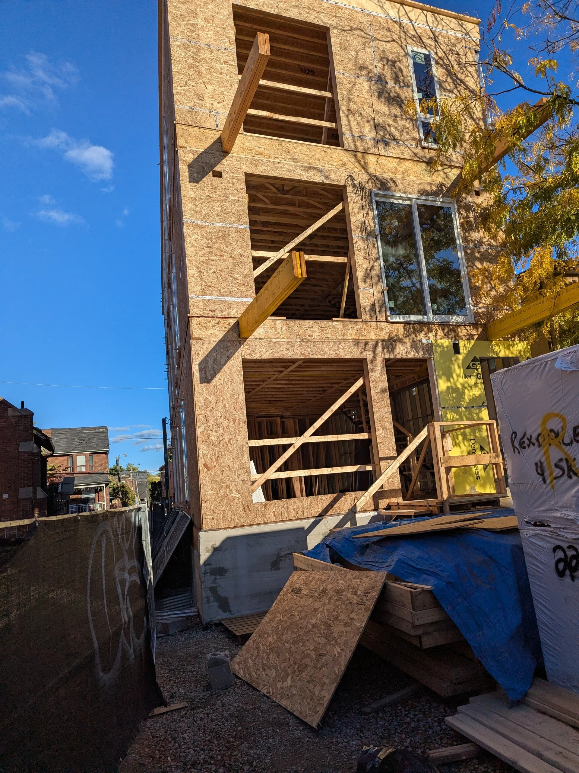 A multi-story building under construction with exposed plywood walls and window frames, scaffolding, and construction materials, including wood and insulation, at a construction site.