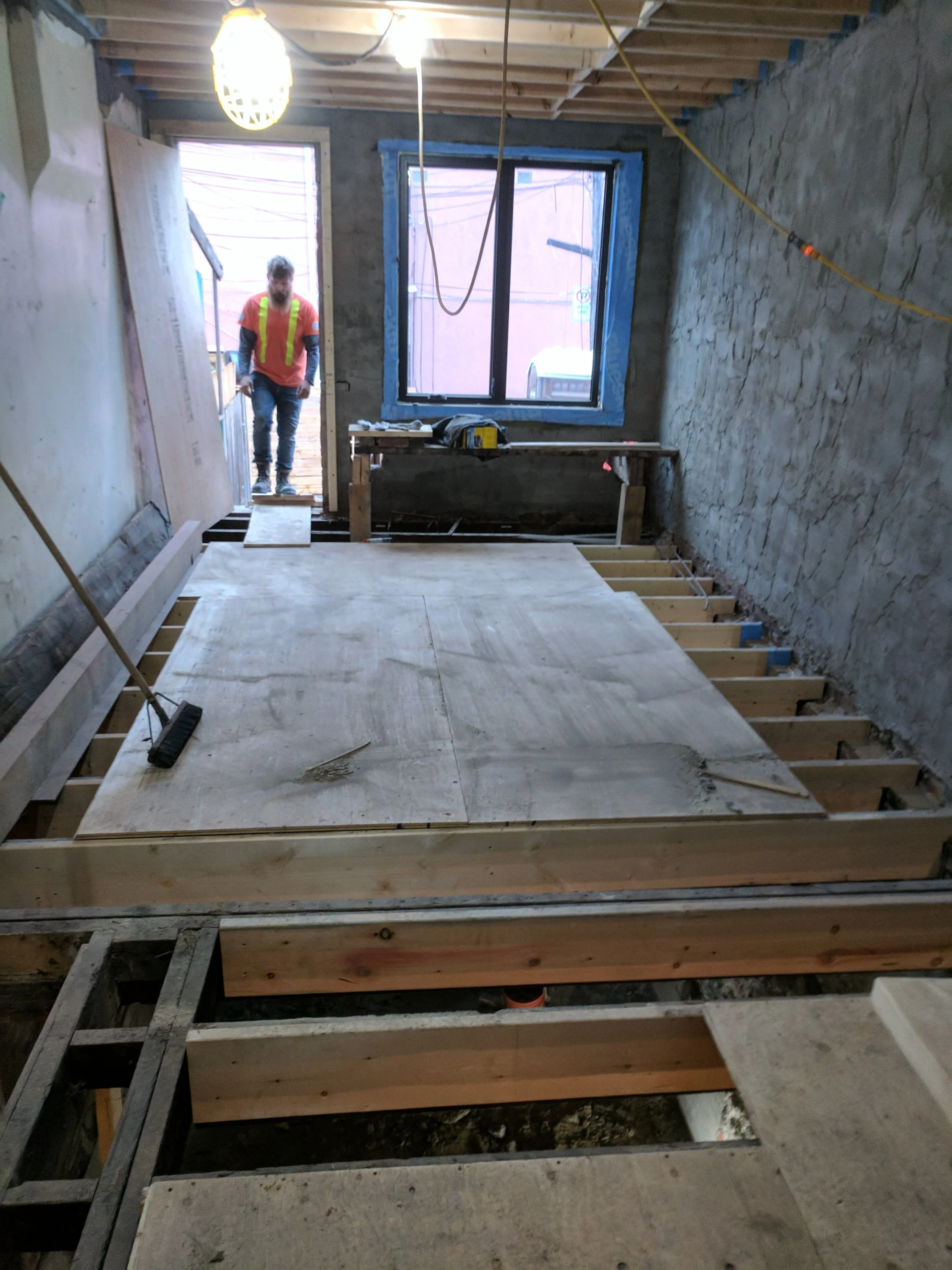 A construction worker in a safety vest standing at a doorway inside a building under renovation, with framing, concrete walls, and a large window.