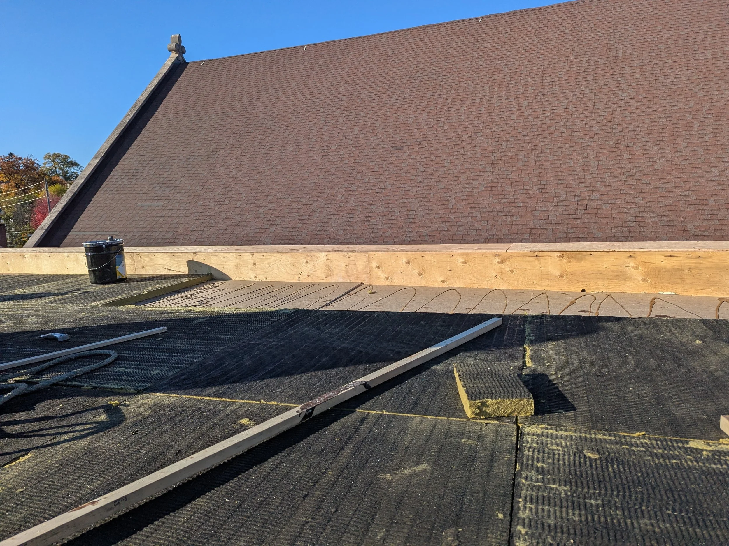 Roof under construction with black waterproofing material, wooden support beam, ladder, and bucket, against a house with a sloped brown shingle roof under a clear blue sky.