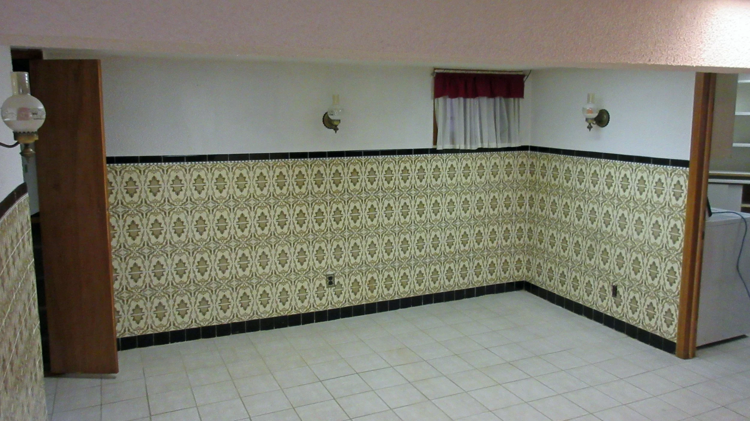 Empty room with patterned yellow and white wallpaper, tiled beige floor, and wood-paneled accents; small window with red and white curtains; wall-mounted lamps.