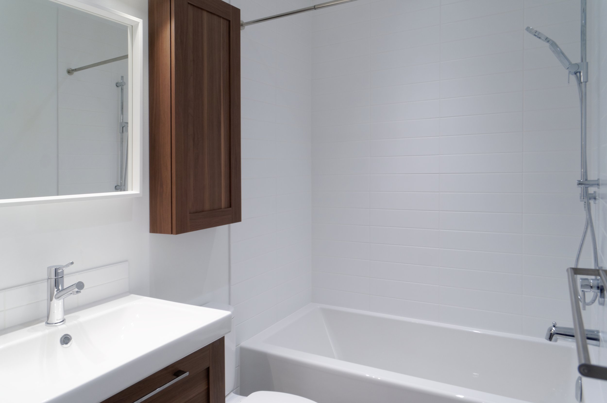 Bathroom with white tiled walls, a white bathtub, a wooden cabinet, a mirror, and a chrome showerhead.