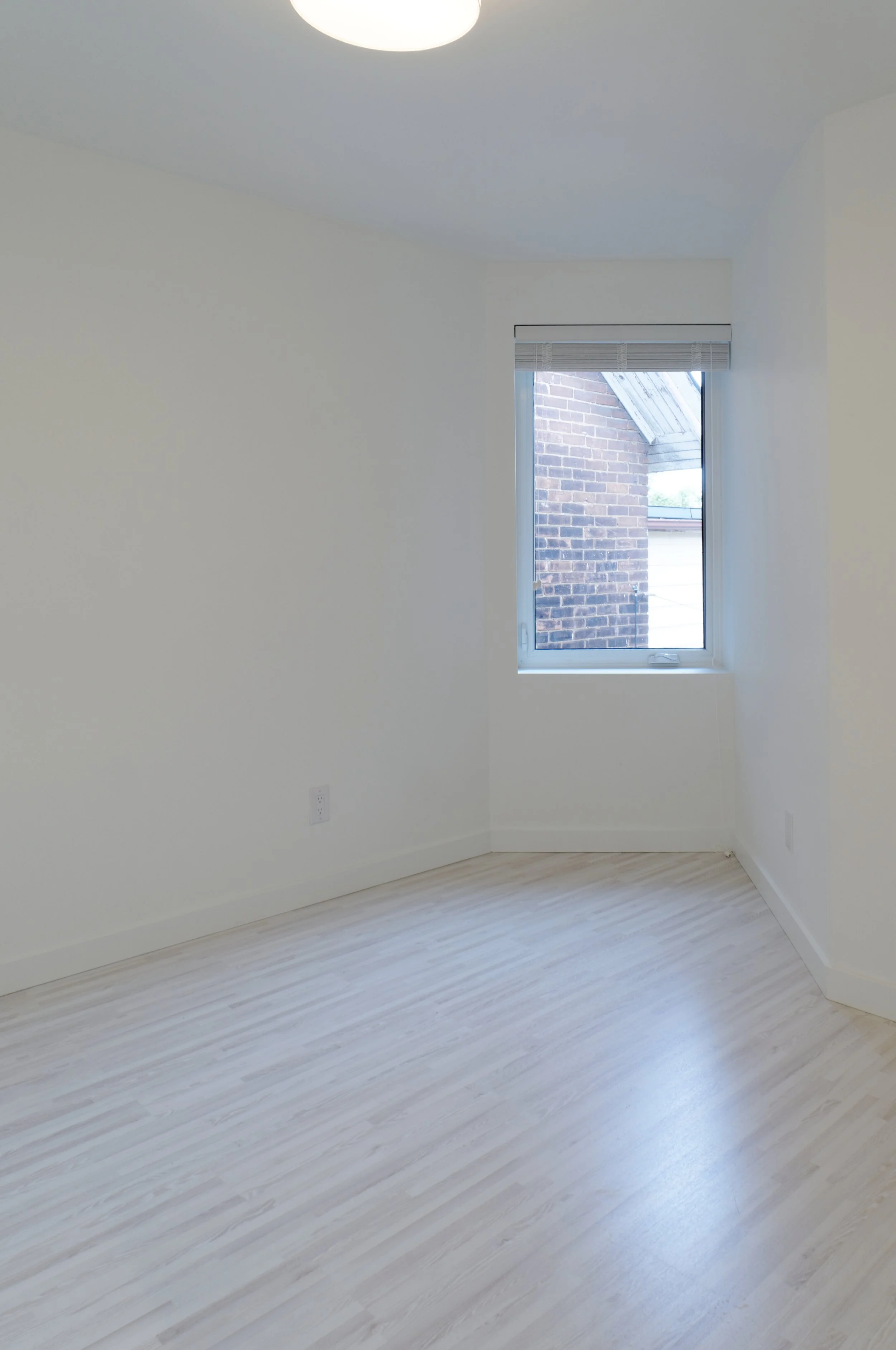 Empty room with white walls, light-colored wooden floor, a window with blinds, and a ceiling light fixture.