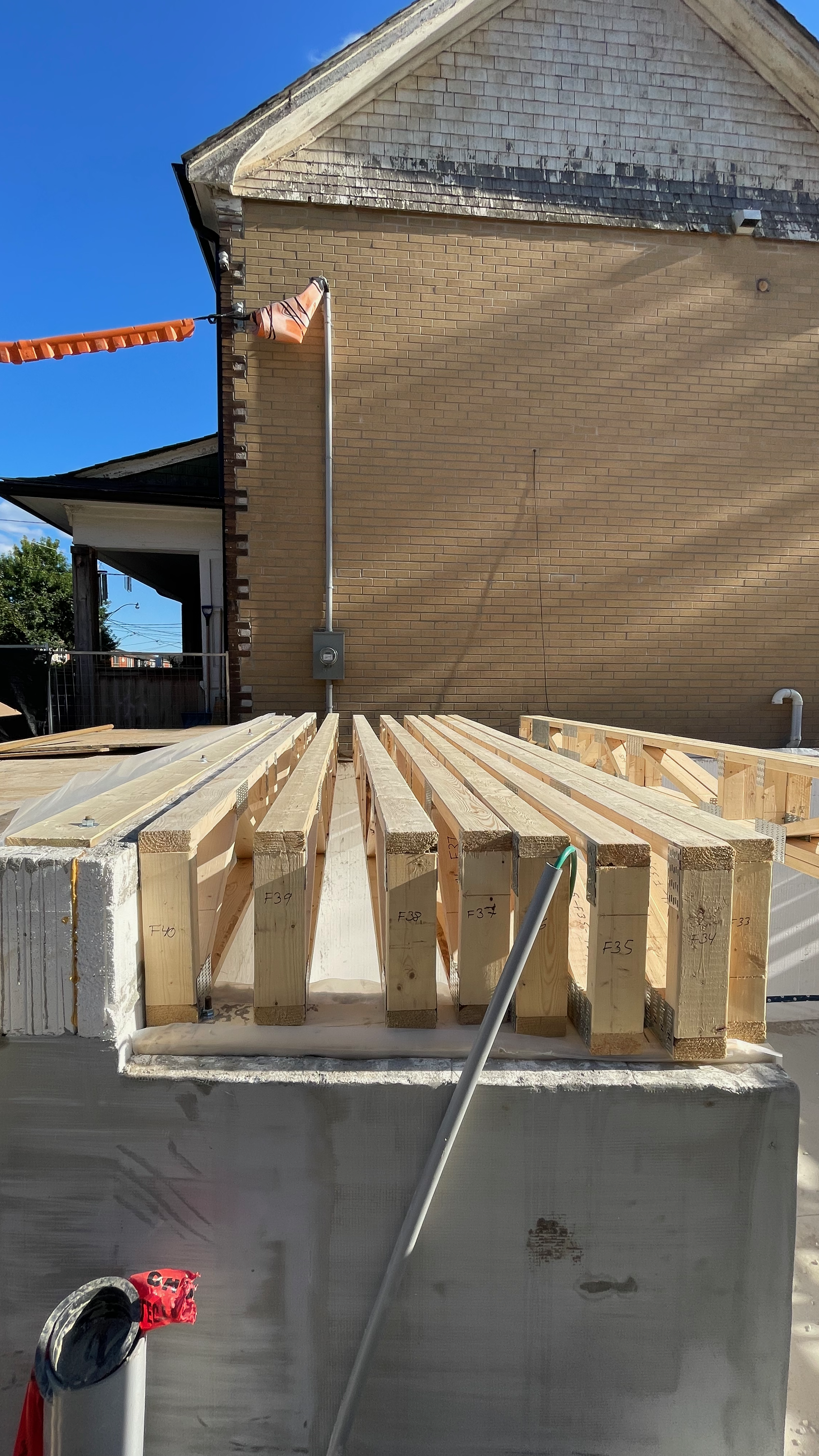 Construction site with wooden framing on top of a concrete foundation, adjacent to a brick house with an electrical box and conduit. A pipe with a green wire is in the foreground, and clear blue sky is overhead.