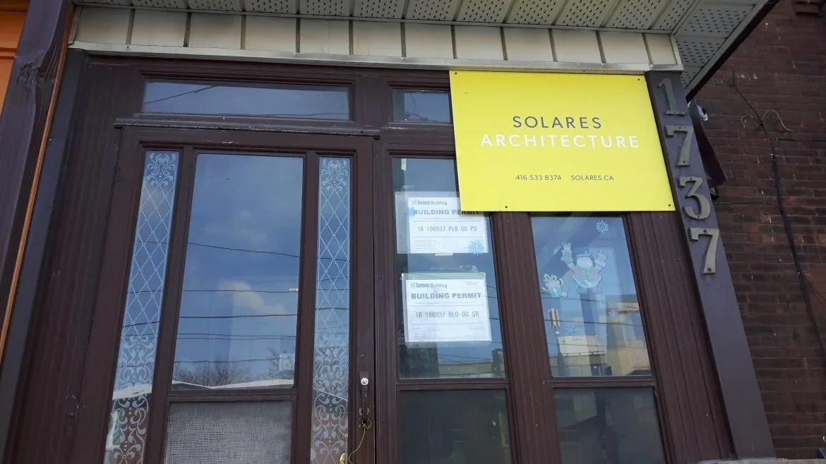Storefront with a yellow sign that reads "Solares Architecture", a phone number, and website, with house numbers 1237 on the side, and window decorations including a snowman sticker and a Santa Claus sticker.