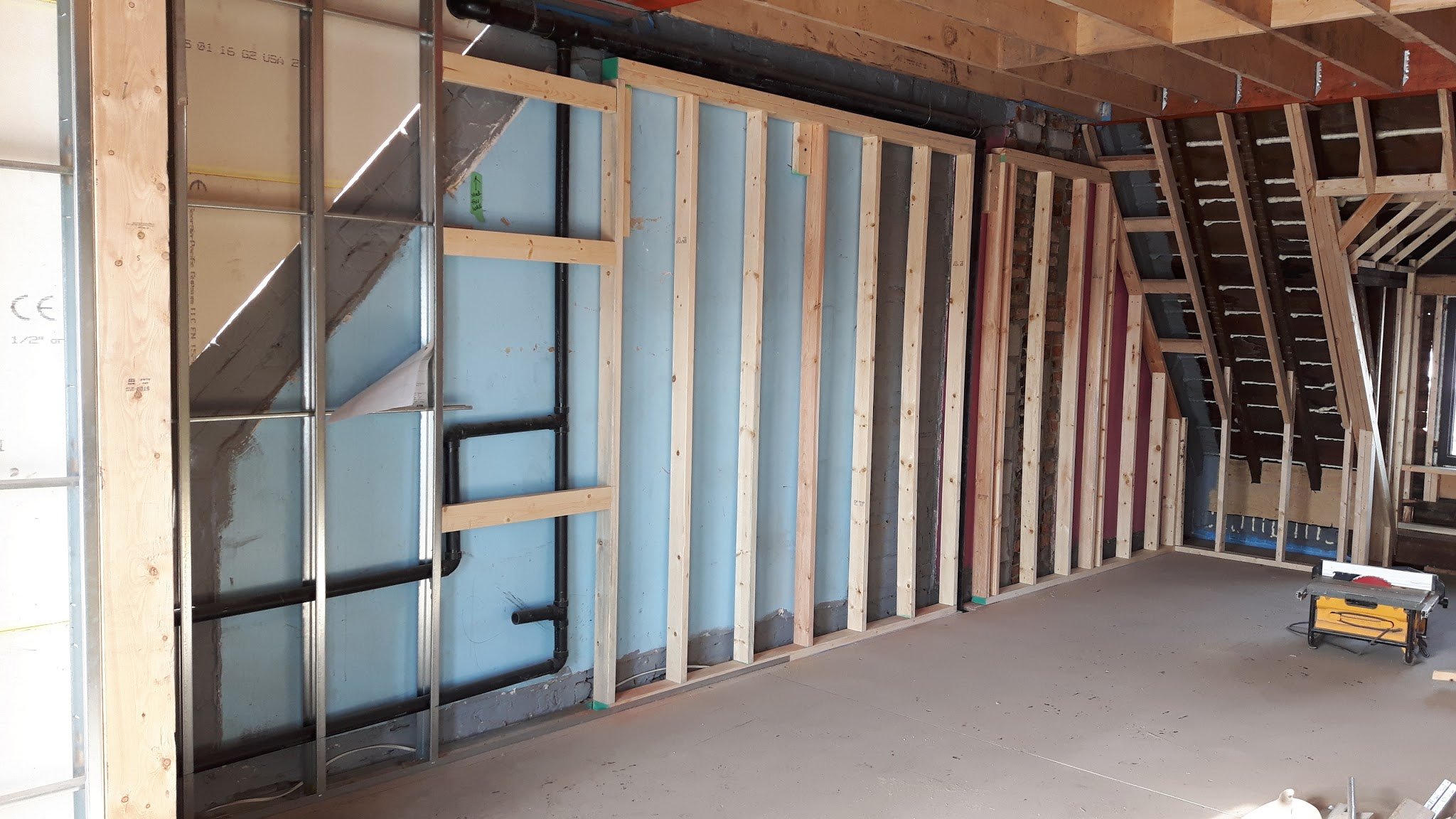 Interior view of a room under construction, showing wooden framing for a wall and ceiling, with visible black plumbing pipes on the wall, and construction tools on the floor.
