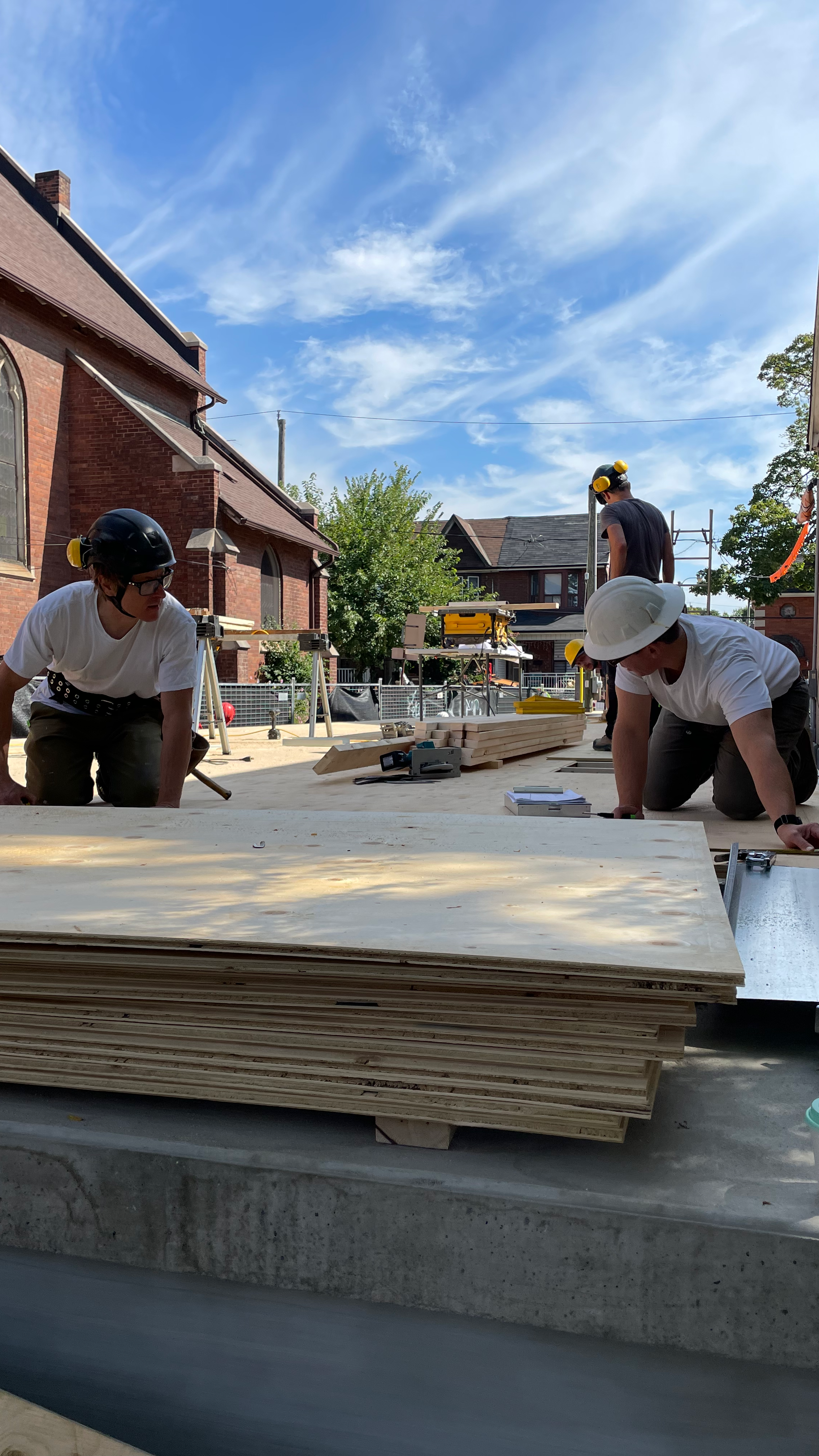 Construction workers wearing safety helmets assemble a floor on a building site with stacks of plywood, power tools, and construction materials, against a background of houses and a blue sky with clouds.