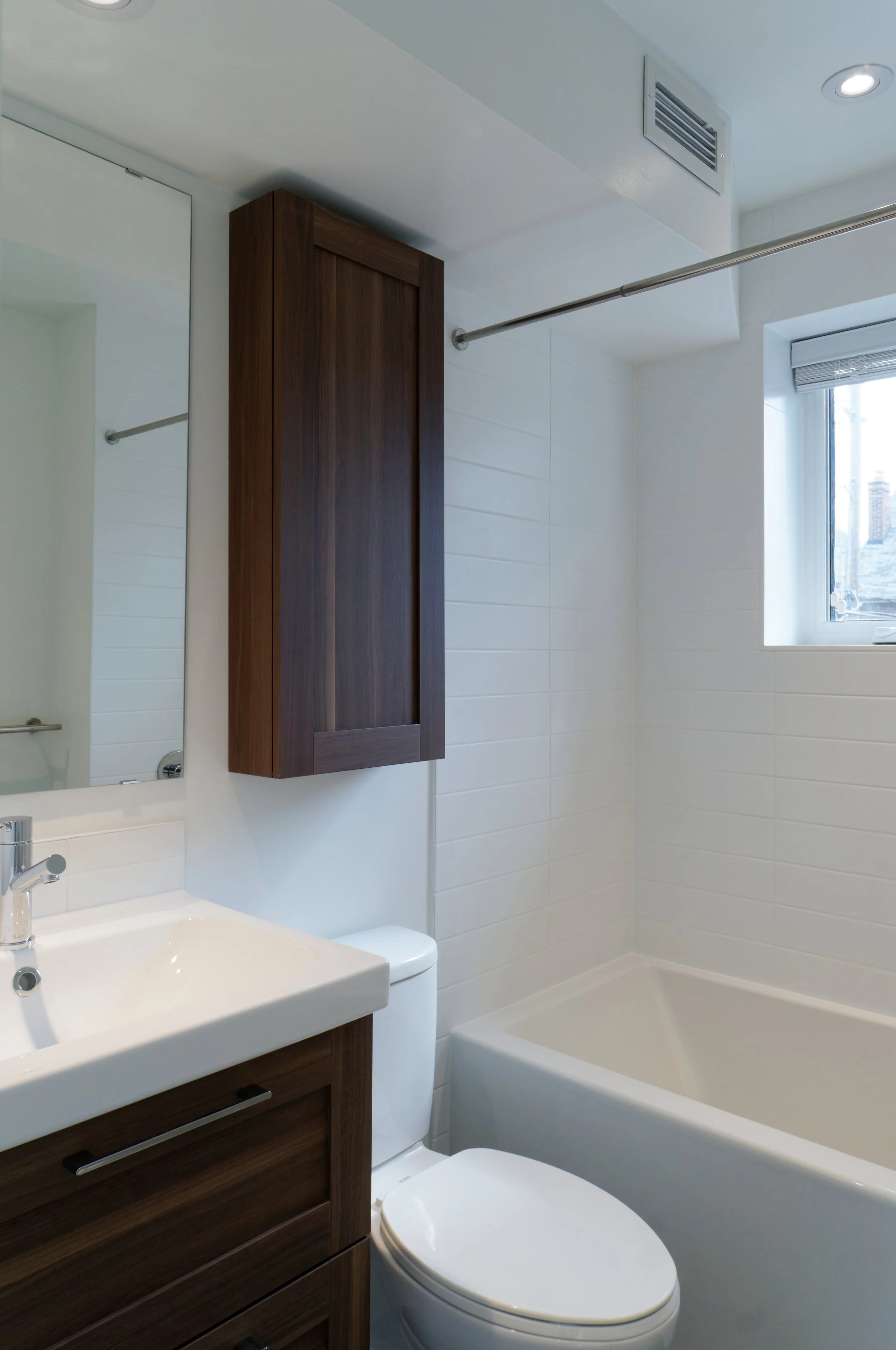 Modern bathroom with white walls, a mirror, a wooden cabinet, a sink with a chrome faucet, a toilet, a bathtub, a window, and a ceiling vent.