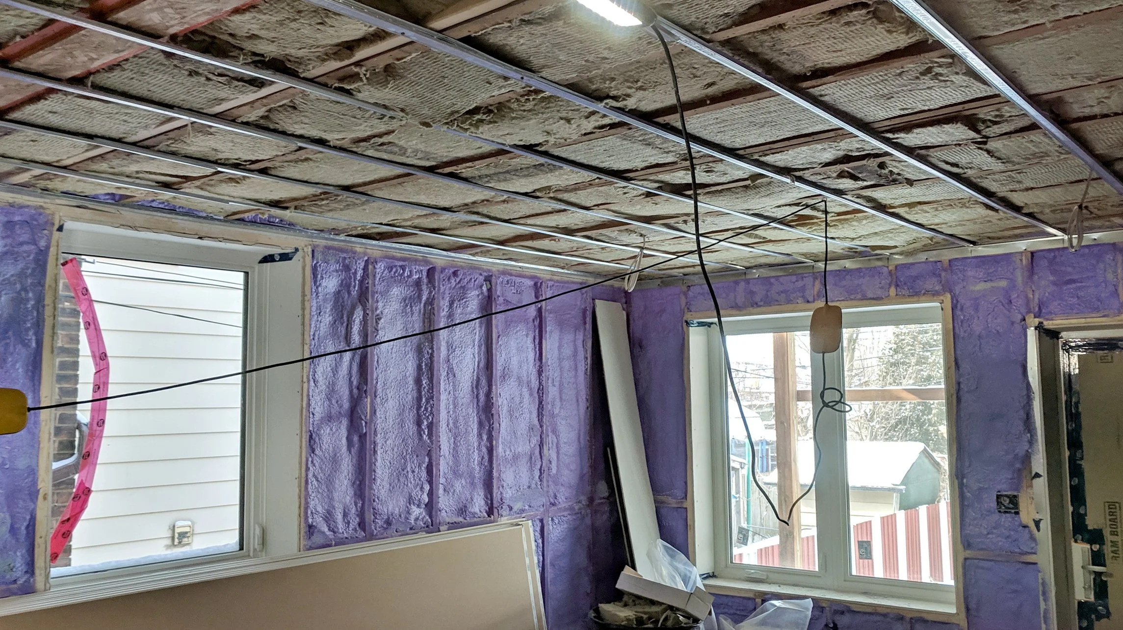 Room under construction with purple insulation on walls, exposed ceiling with insulation and metal framing, and two windows showing snow outside.