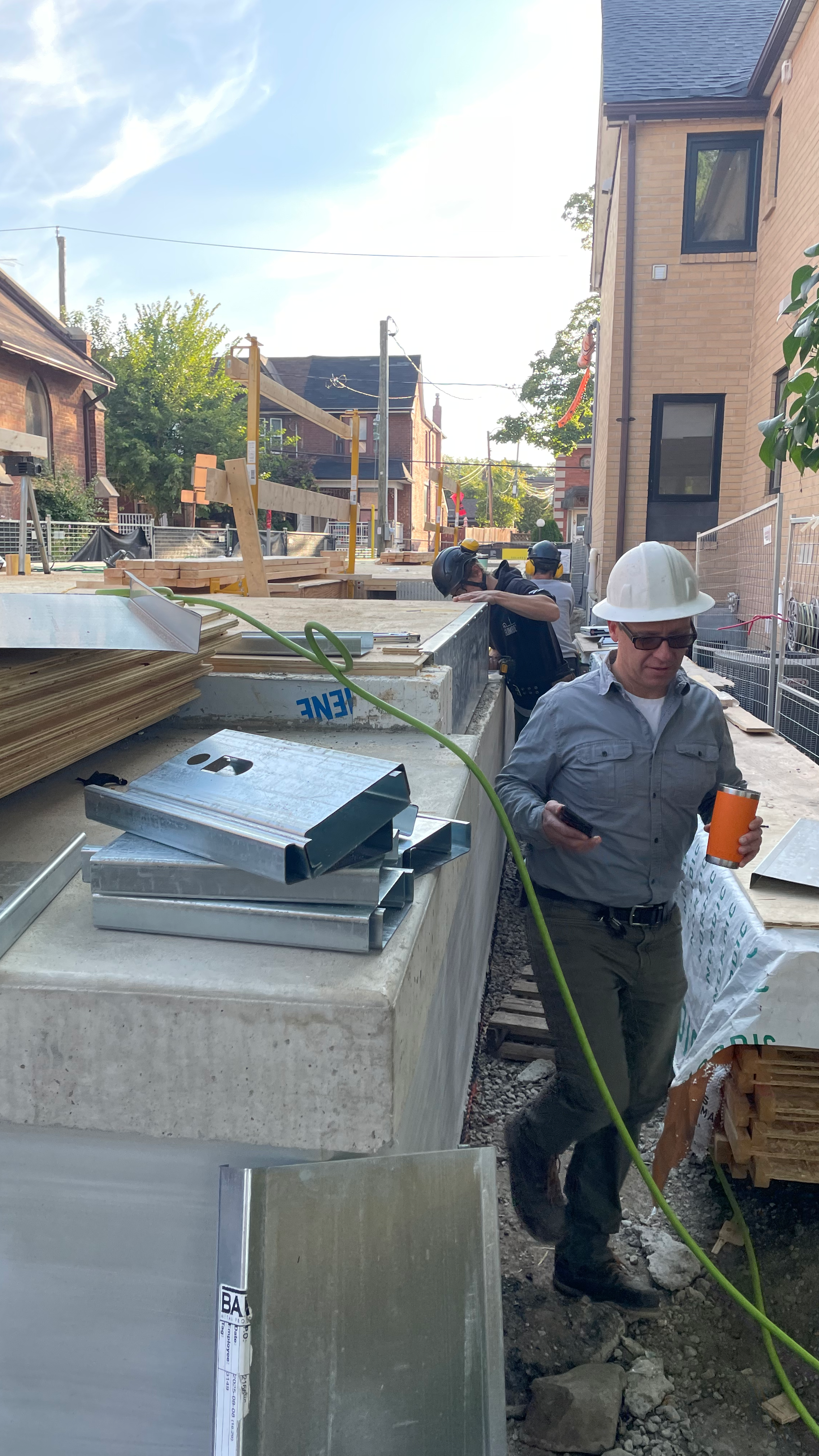 Construction site with workers wearing safety helmets, metal beams, and building materials on a partially built structure in a residential neighborhood.