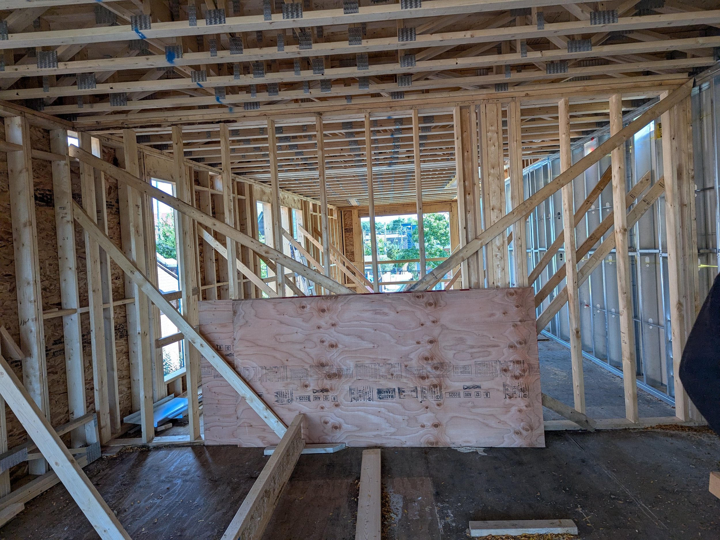 Interior of a house under construction with exposed wooden framing, a plywood temporary wall, and several windows, showing the framework of walls, ceiling, and staircase.