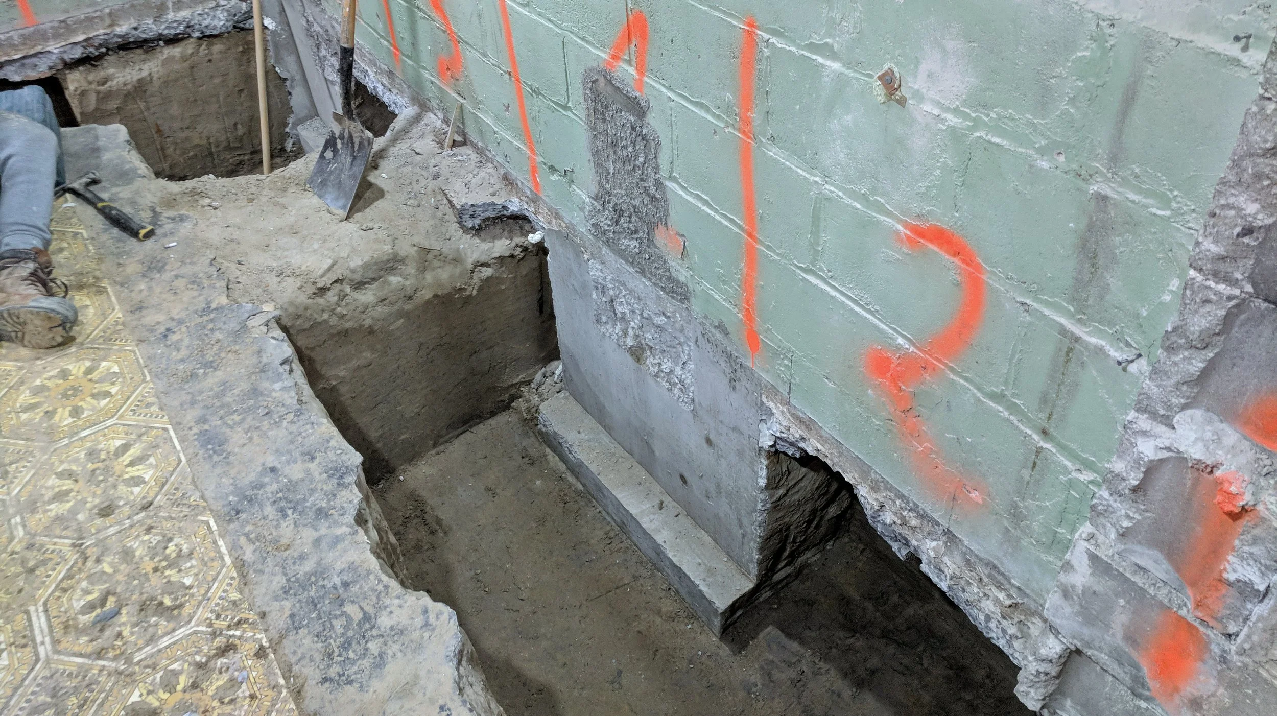 Construction site with a partially excavated area next to a brick wall marked with orange spray paint, construction tools like a hammer and shovel are visible on the ground.