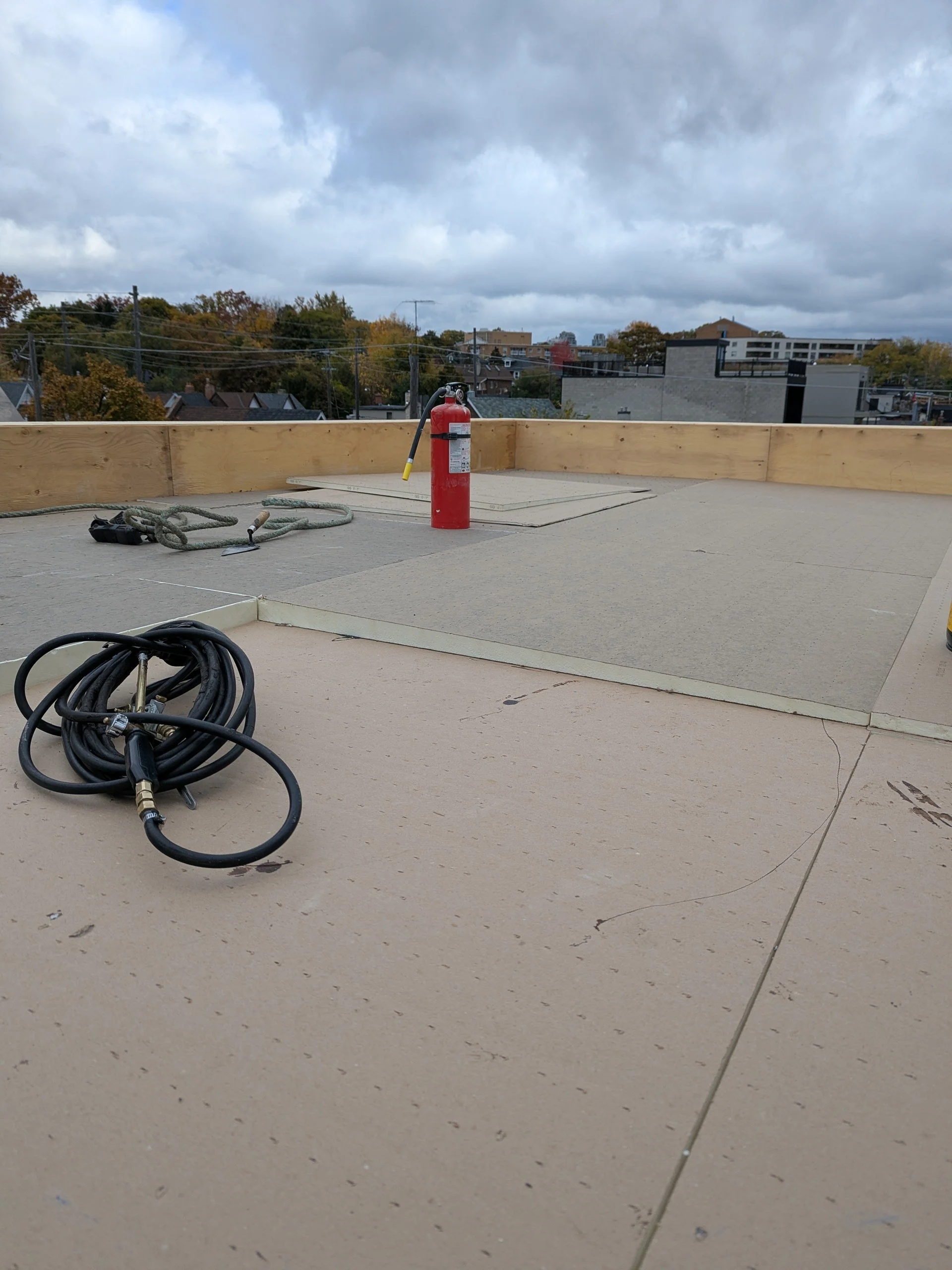 Construction site on a rooftop with a fire extinguisher, ropes, and electrical cables, with city rooftops and cloudy sky in the background.