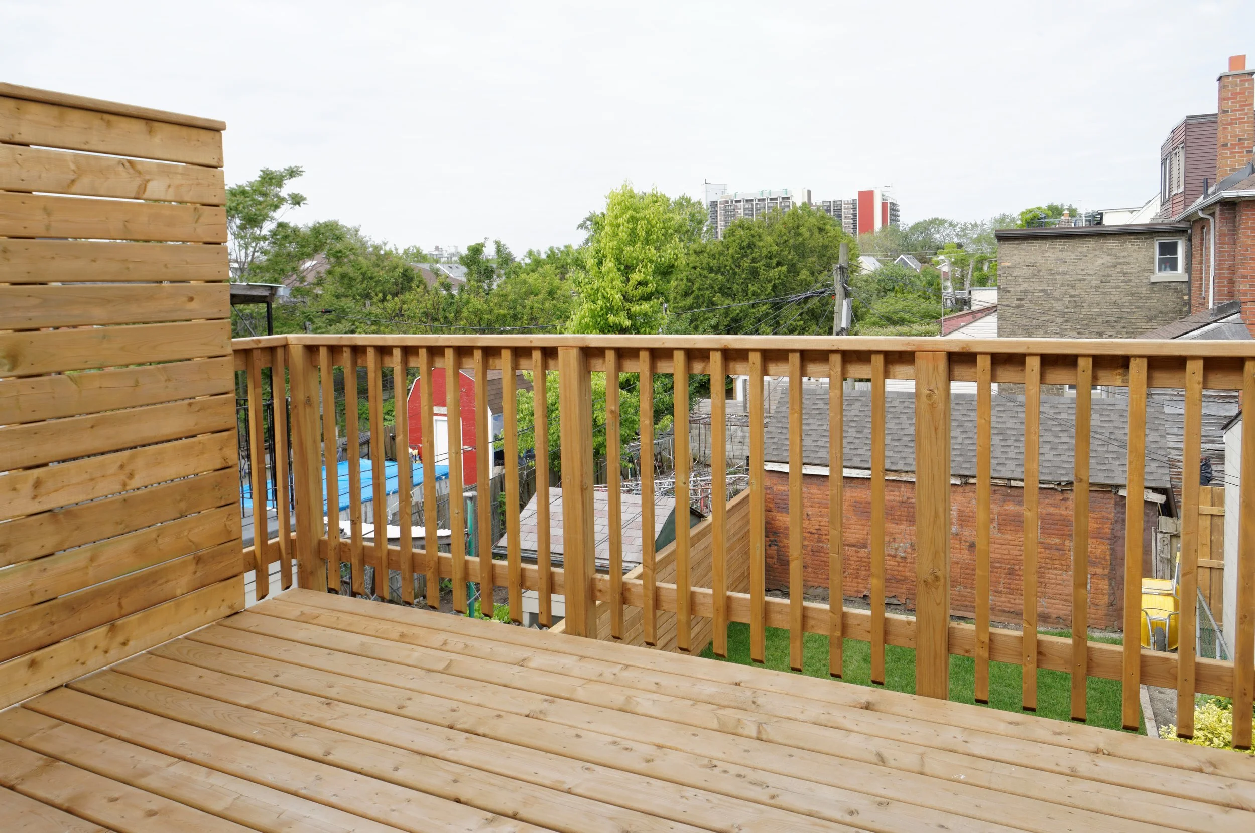 View from a newly built wooden balcony with a railing, overlooking neighboring houses, trees, and distant high-rise buildings under a cloudy sky.