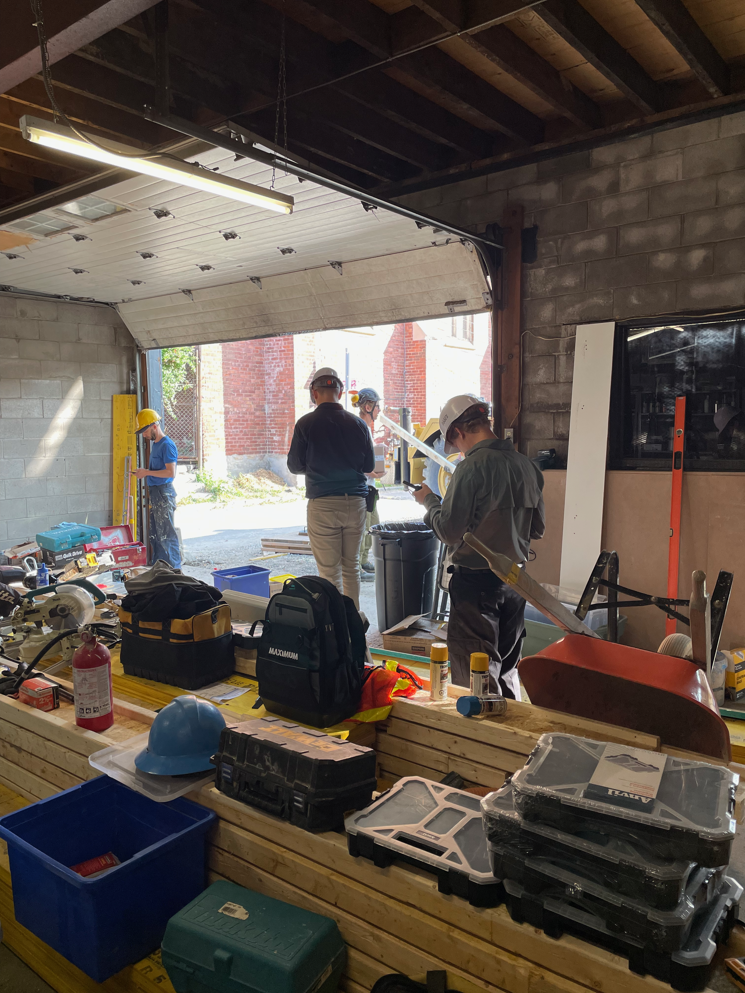 Construction workers and personnel inside a garage or workshop, with tools and equipment on a workbench and outside, some wearing helmets and safety gear.