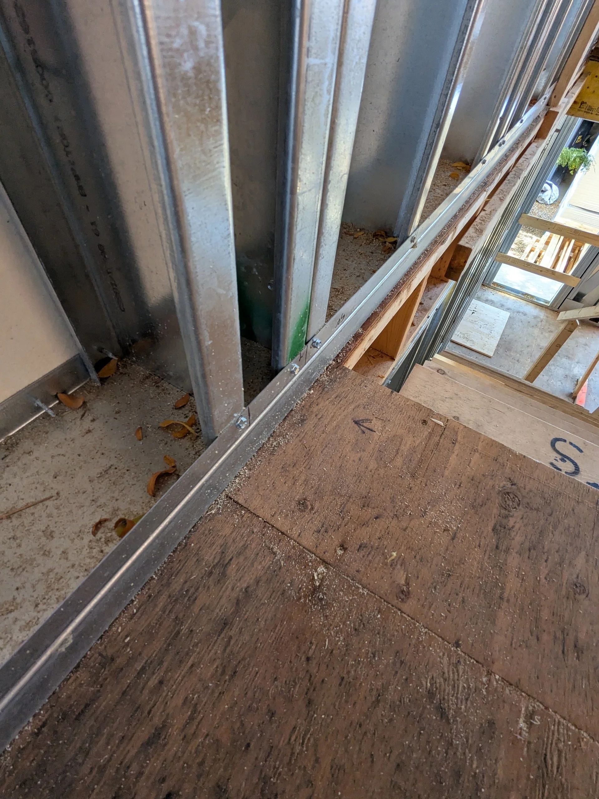 Close-up of a construction site showing metal framing for walls, partially installed alongside wooden flooring, with visible tools, debris, and a partially built staircase in the background.