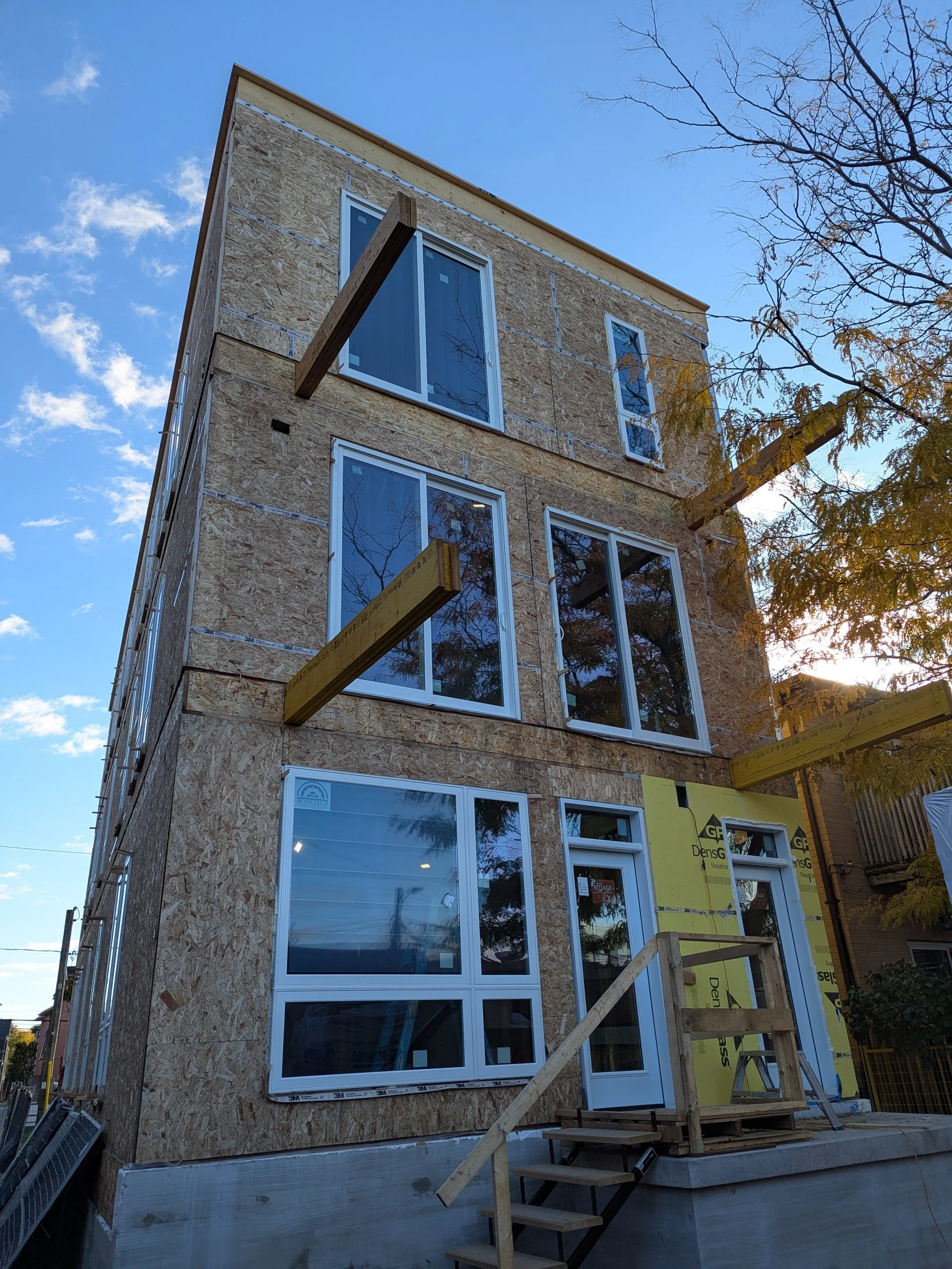 A multi-story building under construction with exterior walls covered in oriented strand board (OSB) sheathing, and windows installed on all floors. The building has a small set of wooden stairs leading up to the front door. The sky is mostly clear w