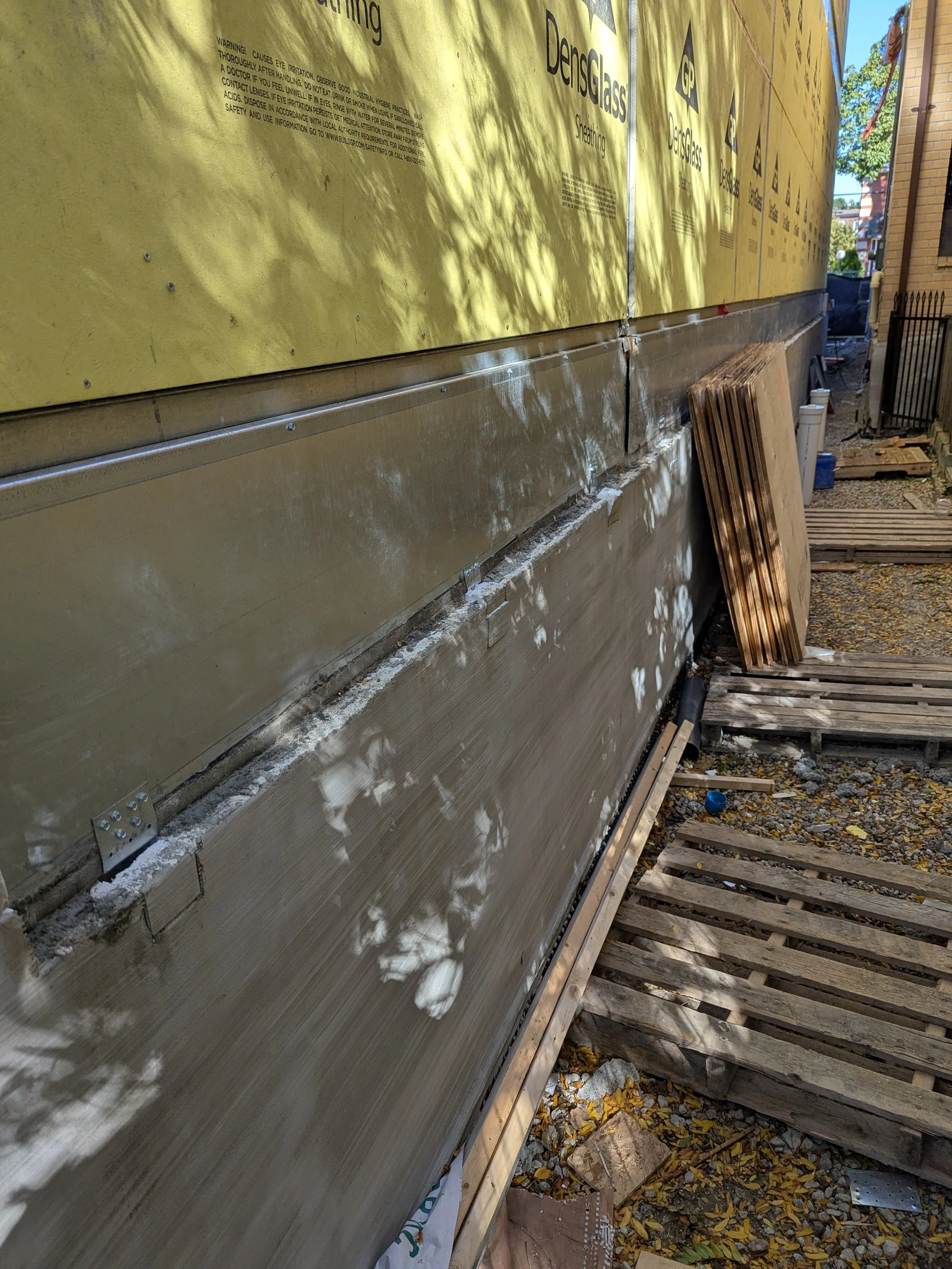 Construction site showing wall insulation with yellow DensGlass sheathing and a metal flashing strip, with wooden pallets and construction materials on the ground.