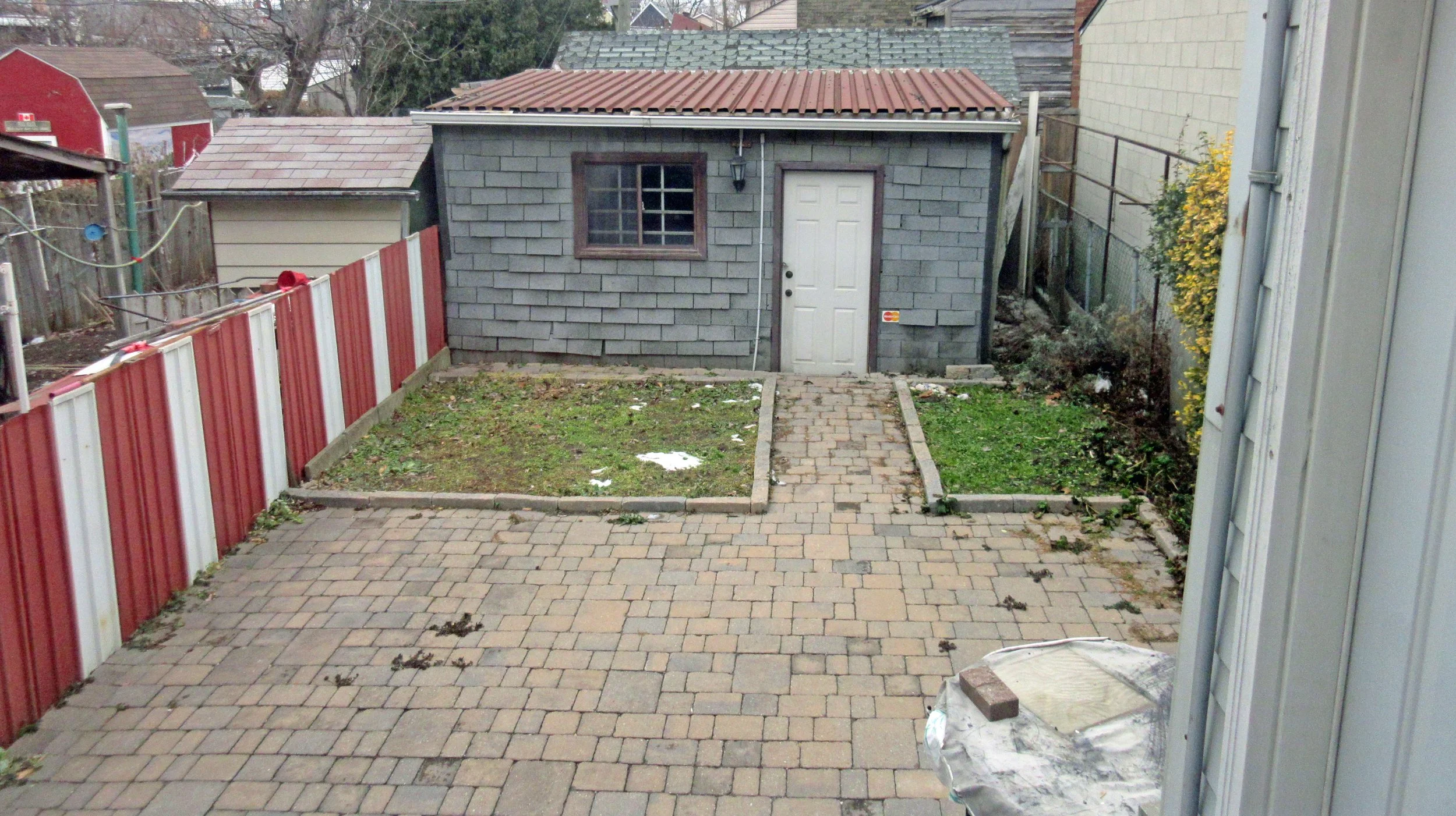 Backyard with a brick patio, small patch of grass, and a gray shed with a white door and a window, bordered by a red and white fence and a black fence on the right. Some scattered debris and patches of dirt are visible.
