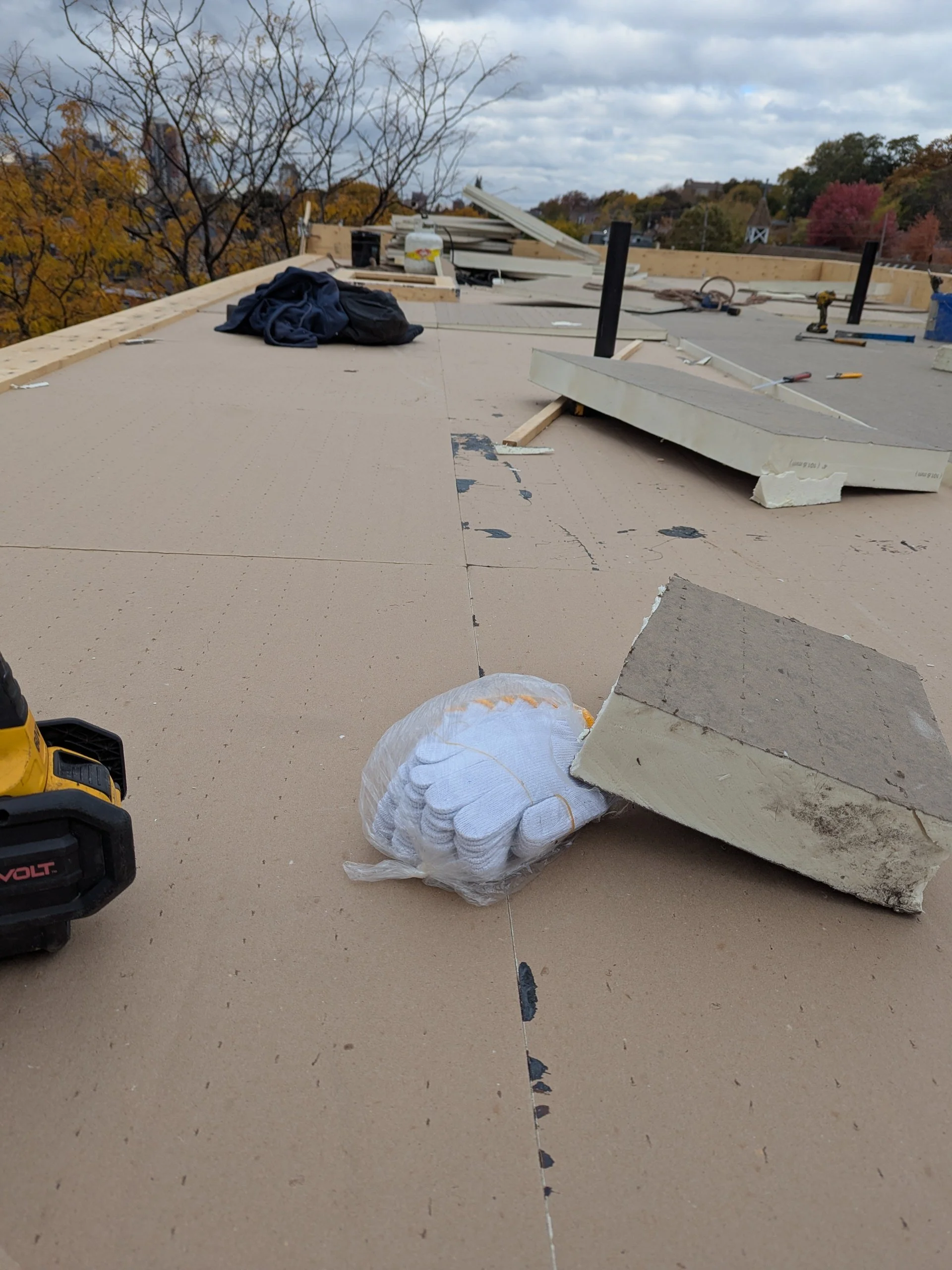 Construction site on a roof with building materials, tools, and a bag of gloves, with trees in the background and overcast sky.