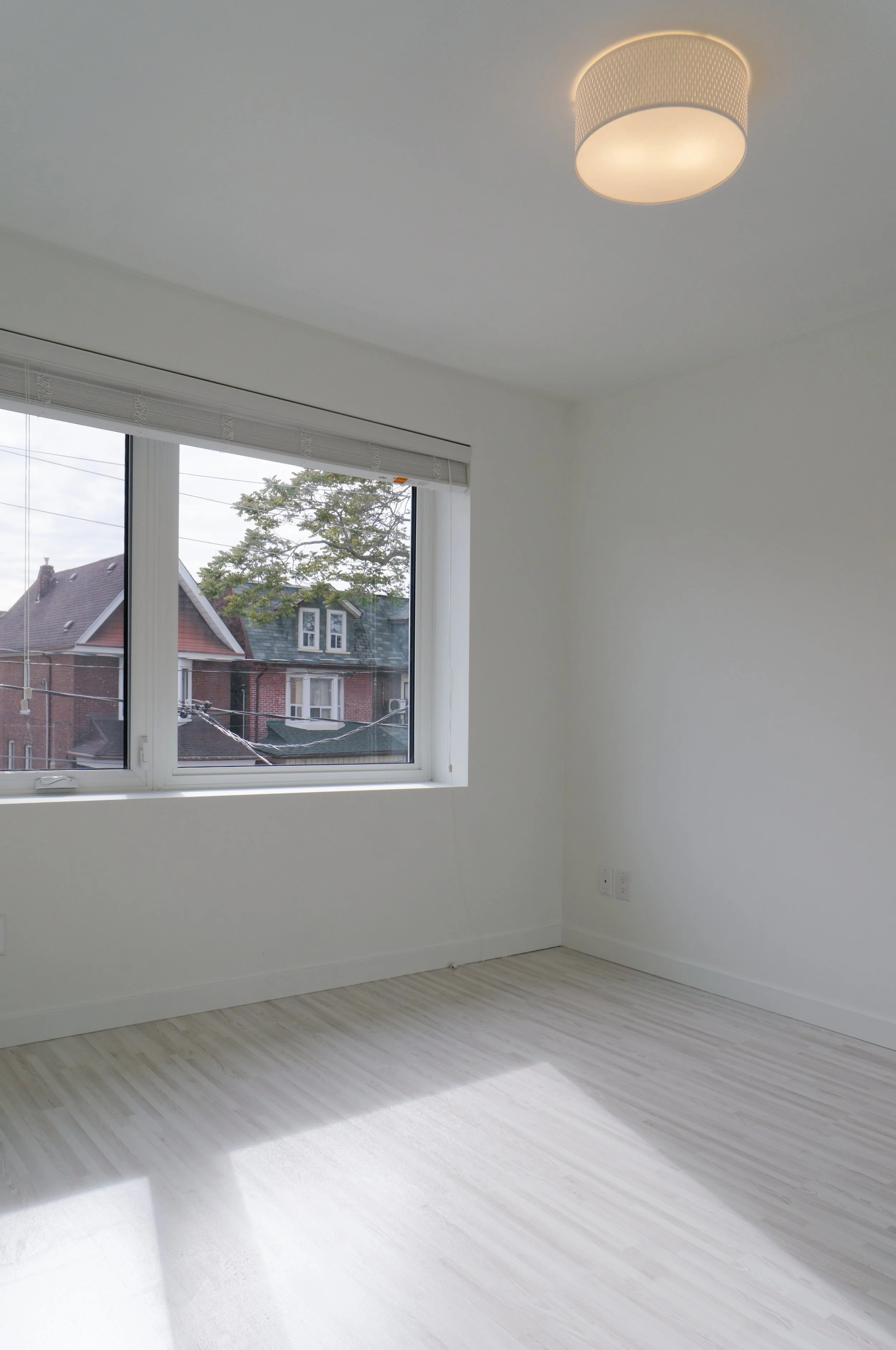 Empty room with white walls, a large window showing neighboring houses, light-colored wooden flooring, and a ceiling light fixture.