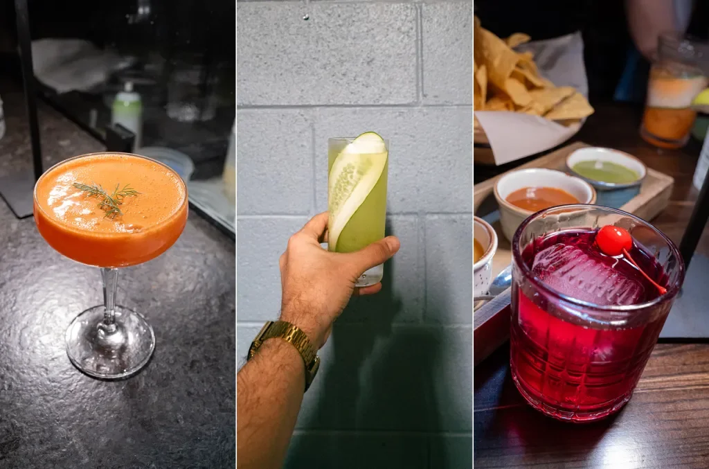 Three cocktails: a mandarin-colored drink with a sprig of rosemary in a coupe glass, a cucumber lime drink with a slice of lime in a tall glass, and a red drink with a cherry on top in a short glass. In the background, there are snacks and sauces.