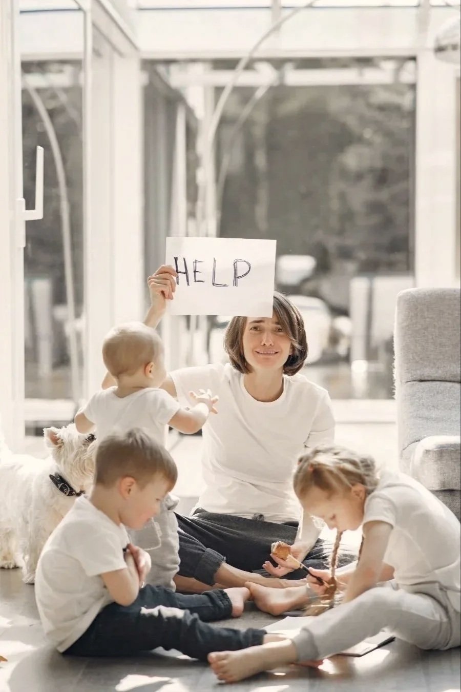 A woman with children and a dog sitting on the floor near a window, holding a sign that says 'HELP', with distressed expressions.