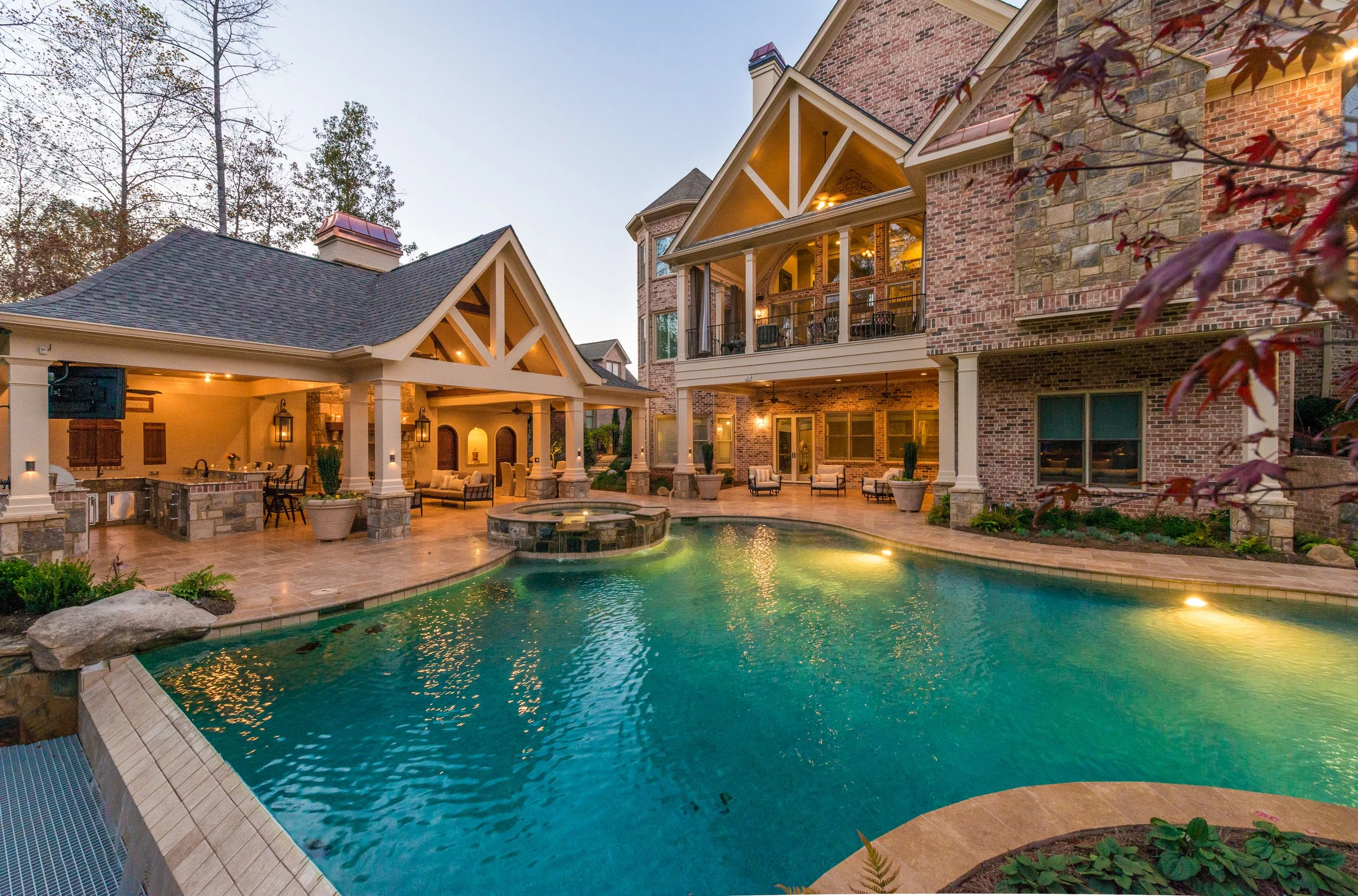 Luxury backyard with a swimming pool, outdoor seating, and a large brick house with multiple stories and balconies, surrounded by trees at dusk.