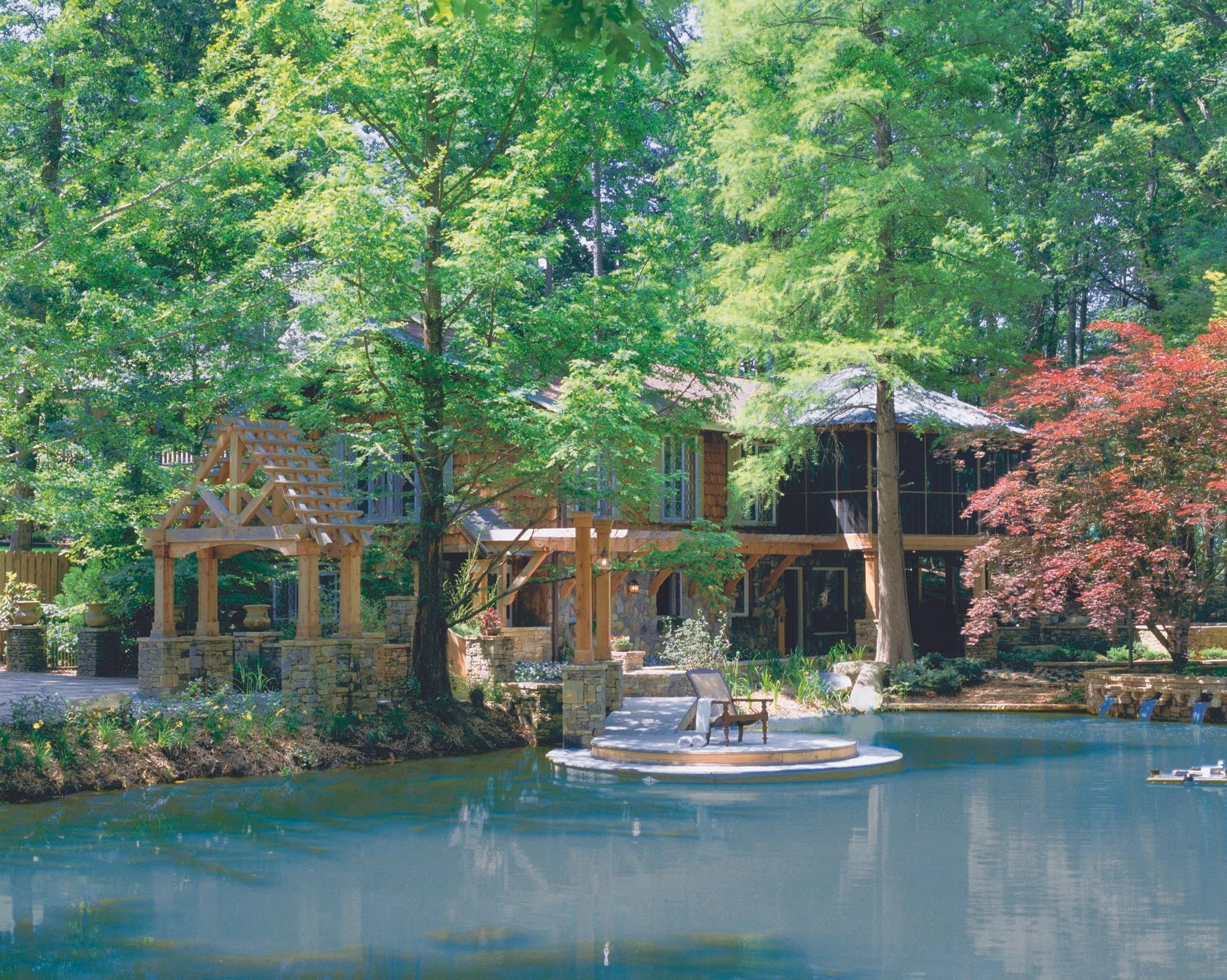 A cozy house surrounded by lush green trees, with a patio and a pond in the foreground.
