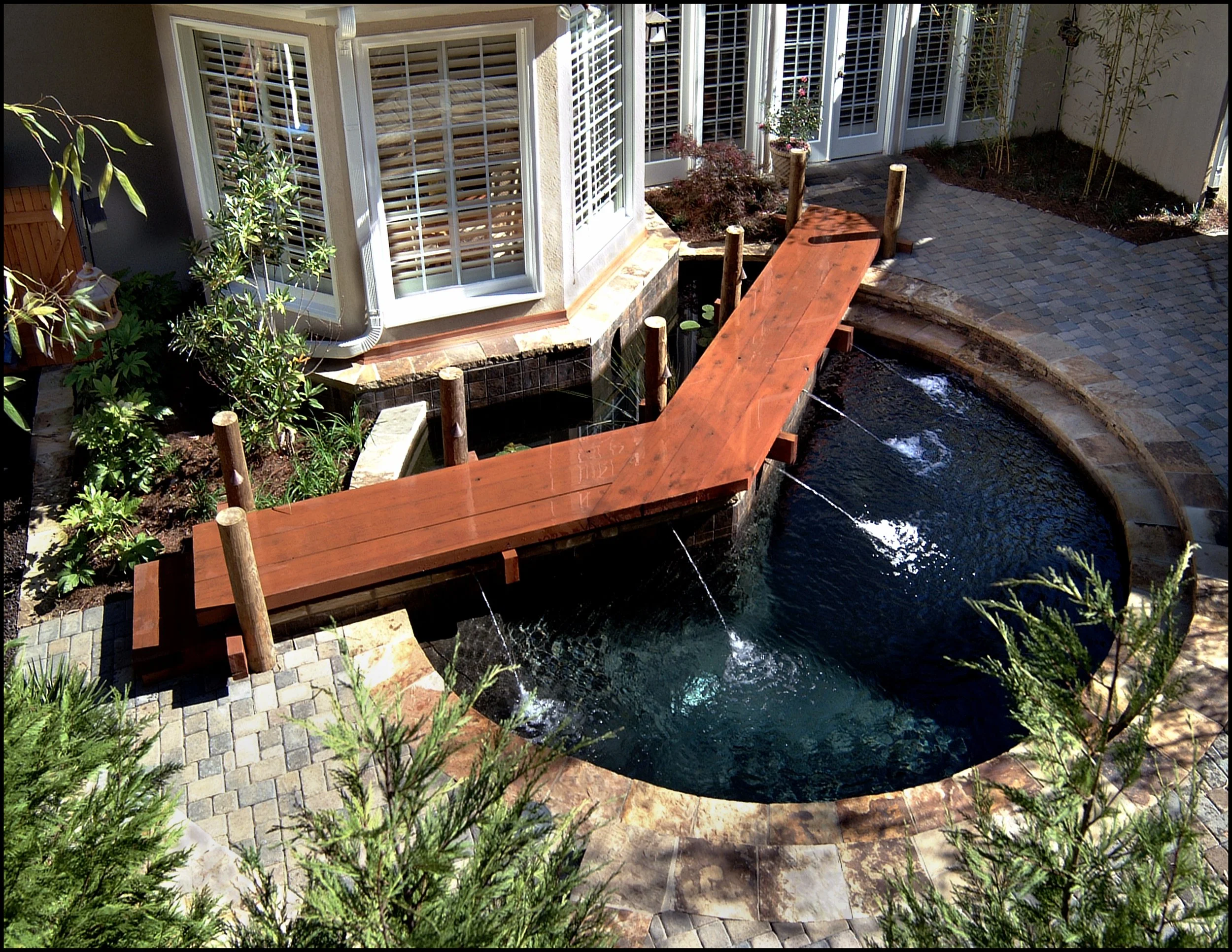Bird's-eye view of a backyard water feature with a curved pond, a small wooden bridge crossing it, surrounded by pavers, plants, and a house with white shutters.