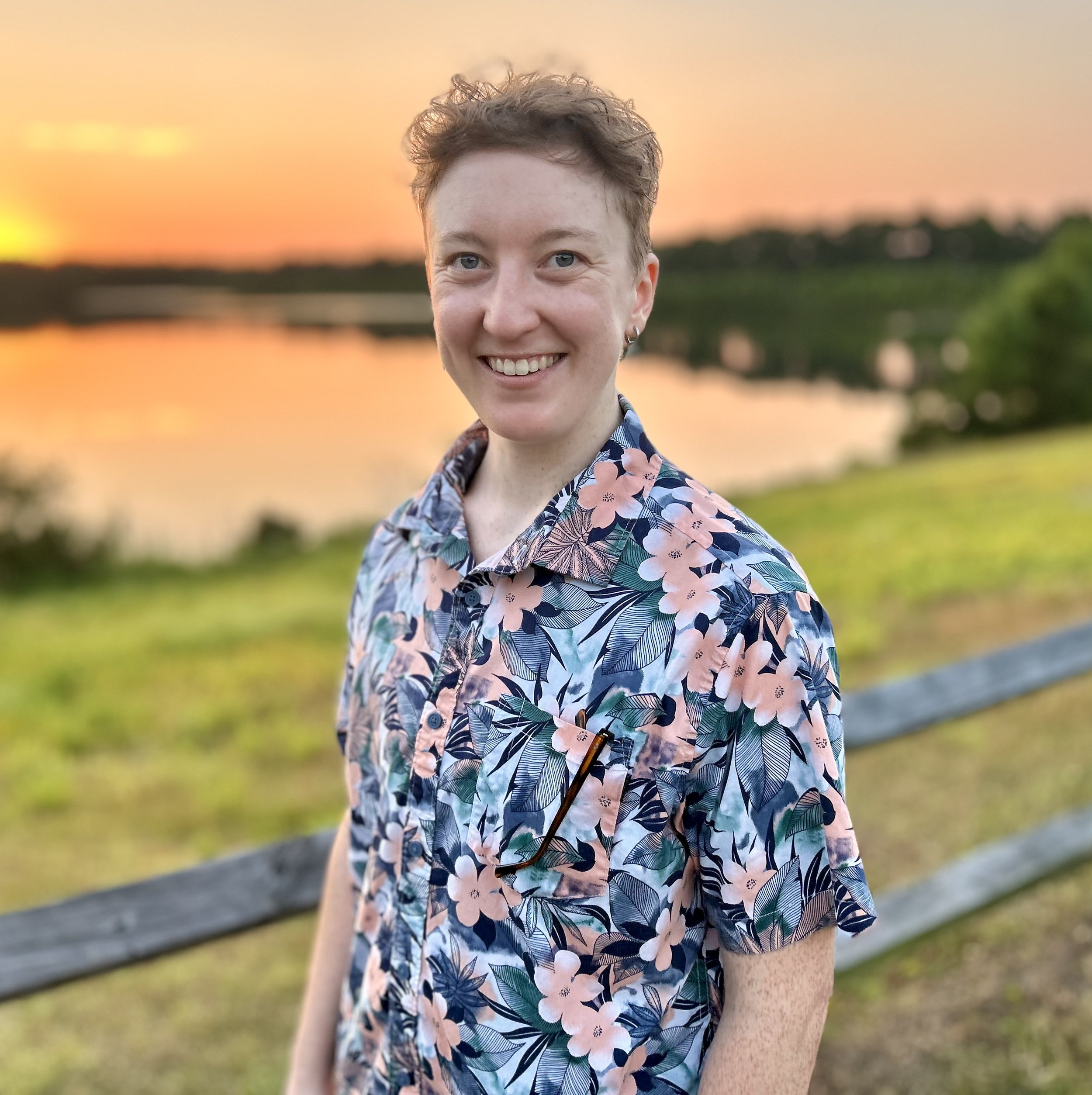 Sam Mitchell, LCHMCA with short brown hair wearing a floral shirt smiling outdoors at sunset by a river.