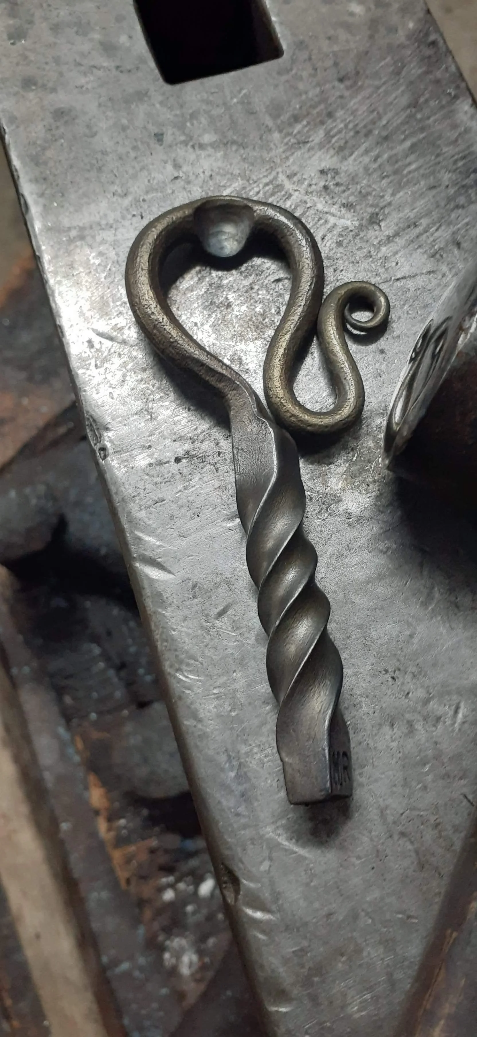 A blacksmithing hammer and a decorative metal hairpin with a spiral and snake design on a metal workbench.