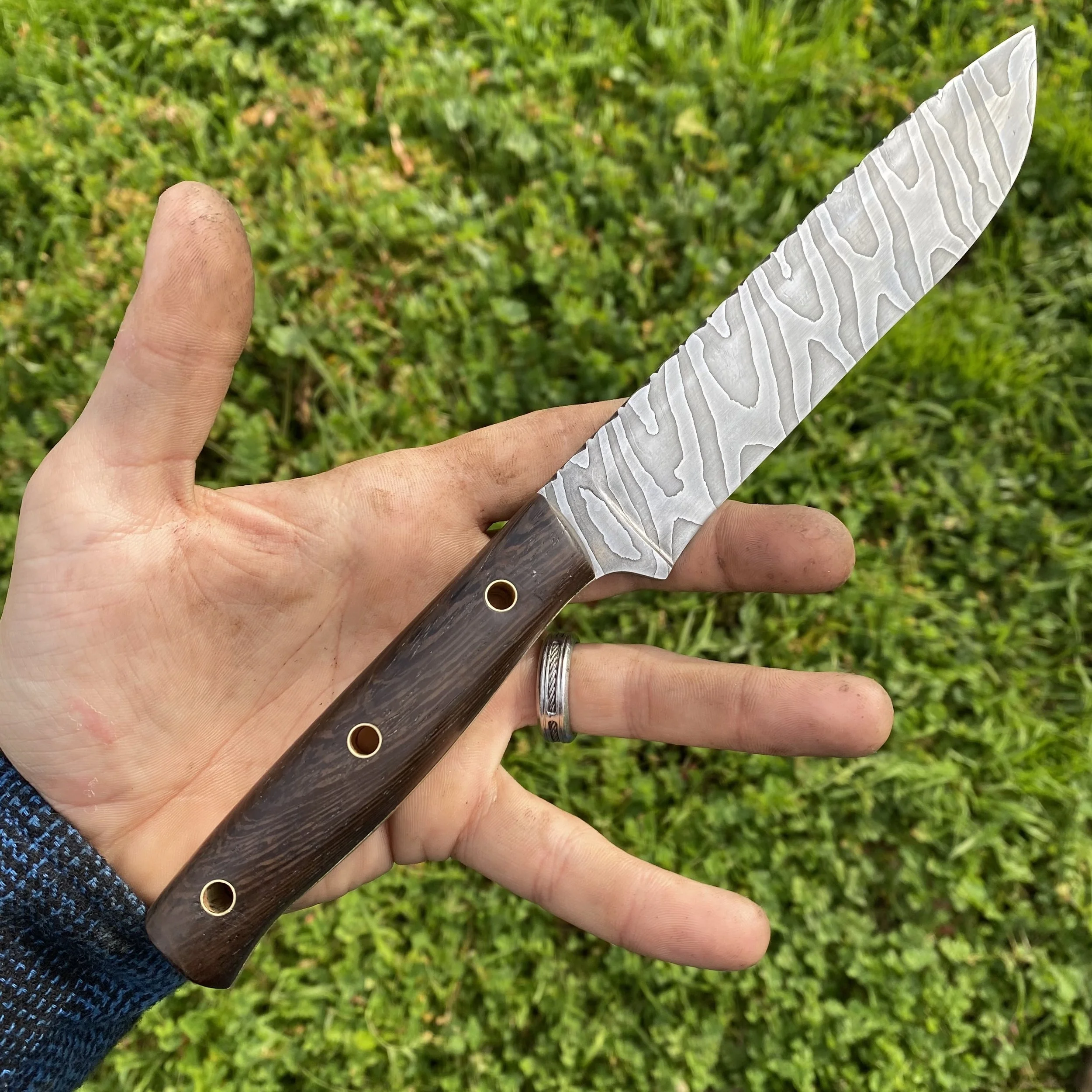 Hand holding a Damascus steel knife with a wooden handle, outdoors on green grass.