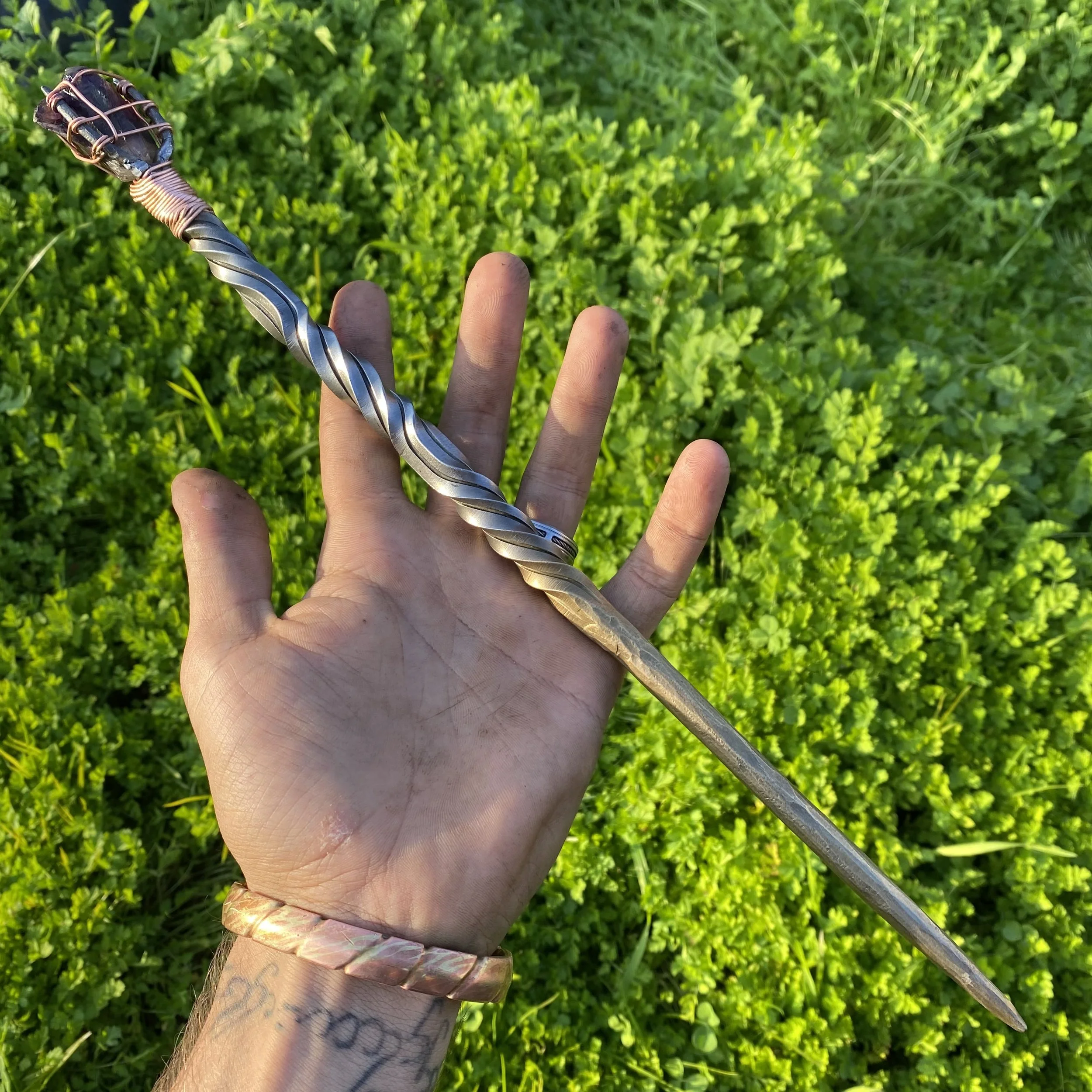 Hand holding a large metallic twisted drill bit over green grass.