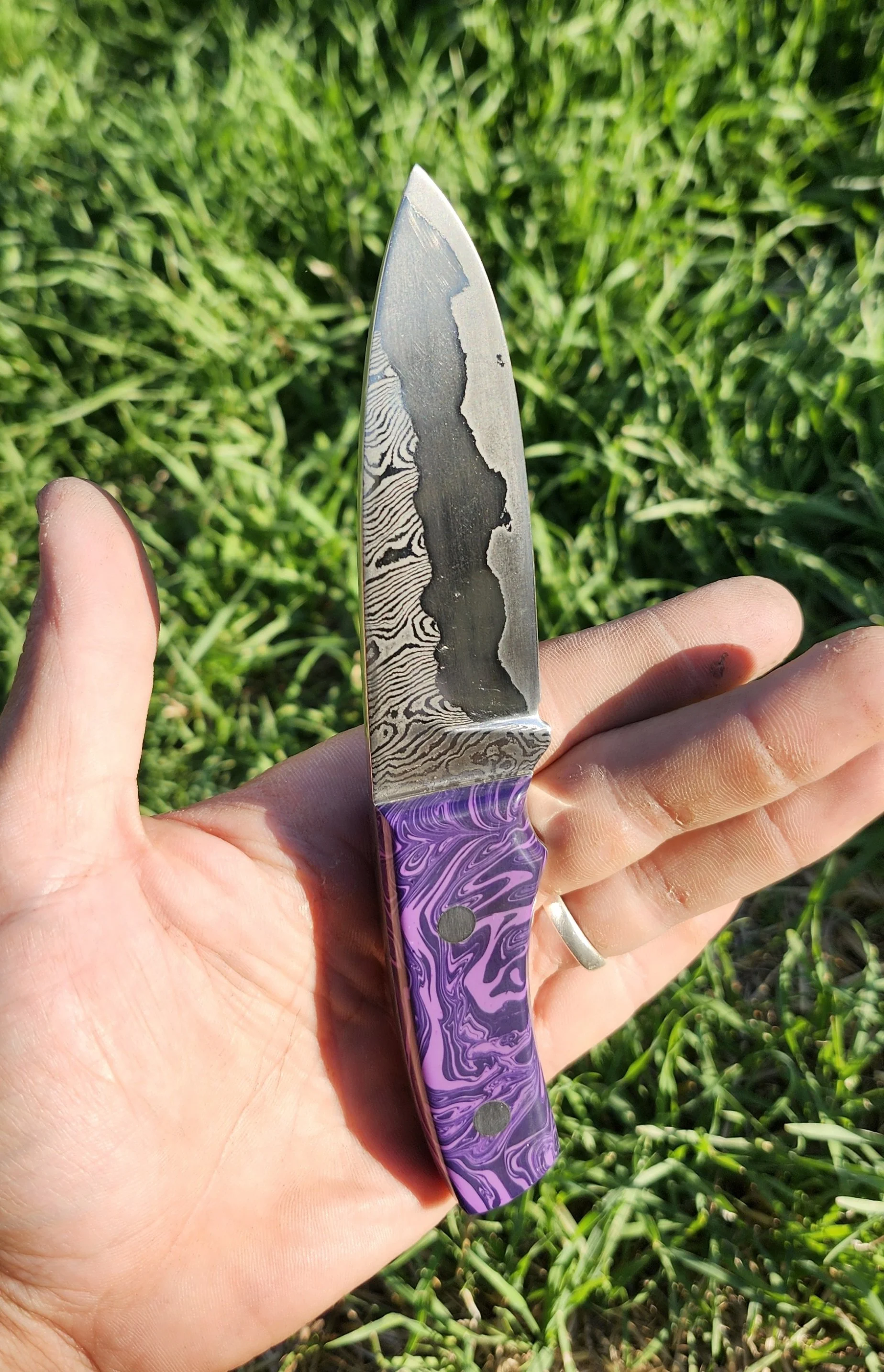 A hand holding a decorative pocket knife with a purple and black marbled handle and a Damascus steel blade, outdoors on green grass.