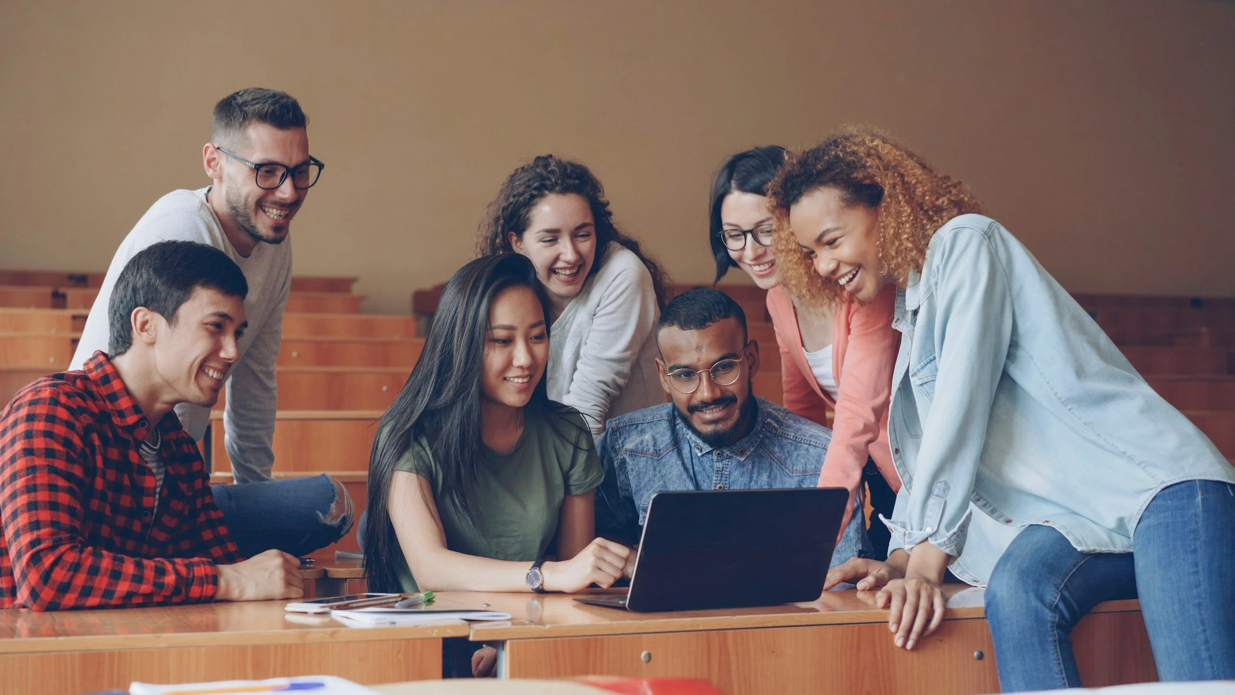 A group of eight diverse young adults gathered around a laptop in a classroom, smiling and looking at the screen together.