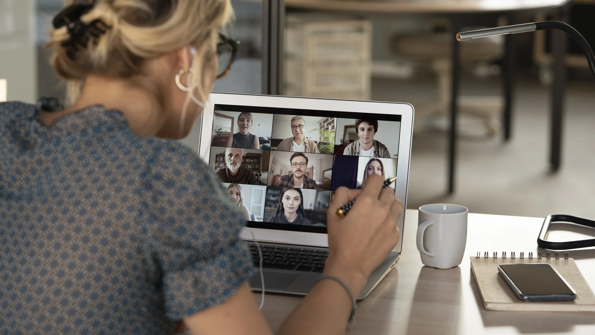 A woman participating in a video conference on her laptop, showing multiple people on the screen, with a notebook, pencil, and smartphone on her desk.