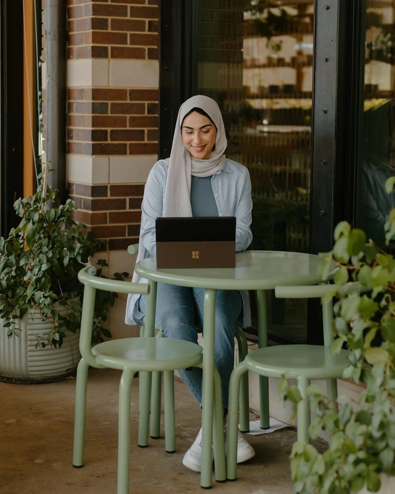 A woman sitting at a green outdoor table using a black laptop next to a potted plant. She has a light gray headscarf and is smiling.