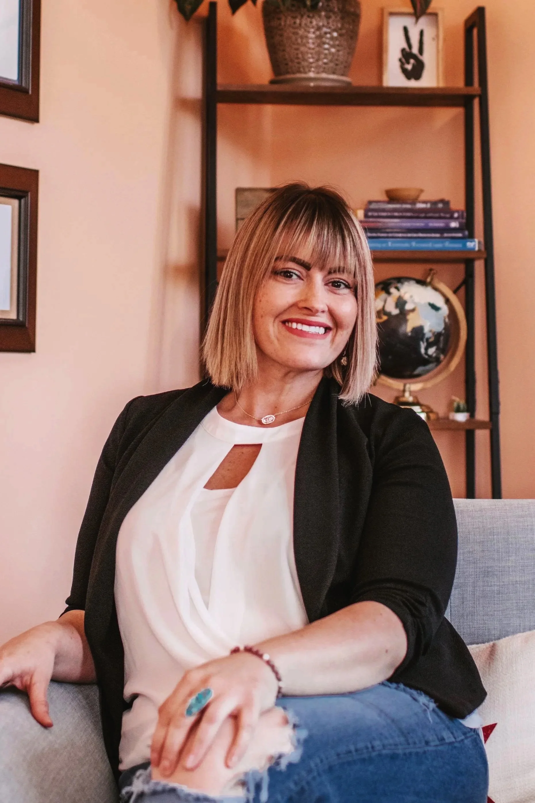 A woman with shoulder-length blonde hair and bangs, smiling, sitting on a couch. She is wearing a black blazer over a white blouse with a keyhole cutout, and blue ripped jeans. Behind her is a bookshelf with books, a globe, and decorative items.