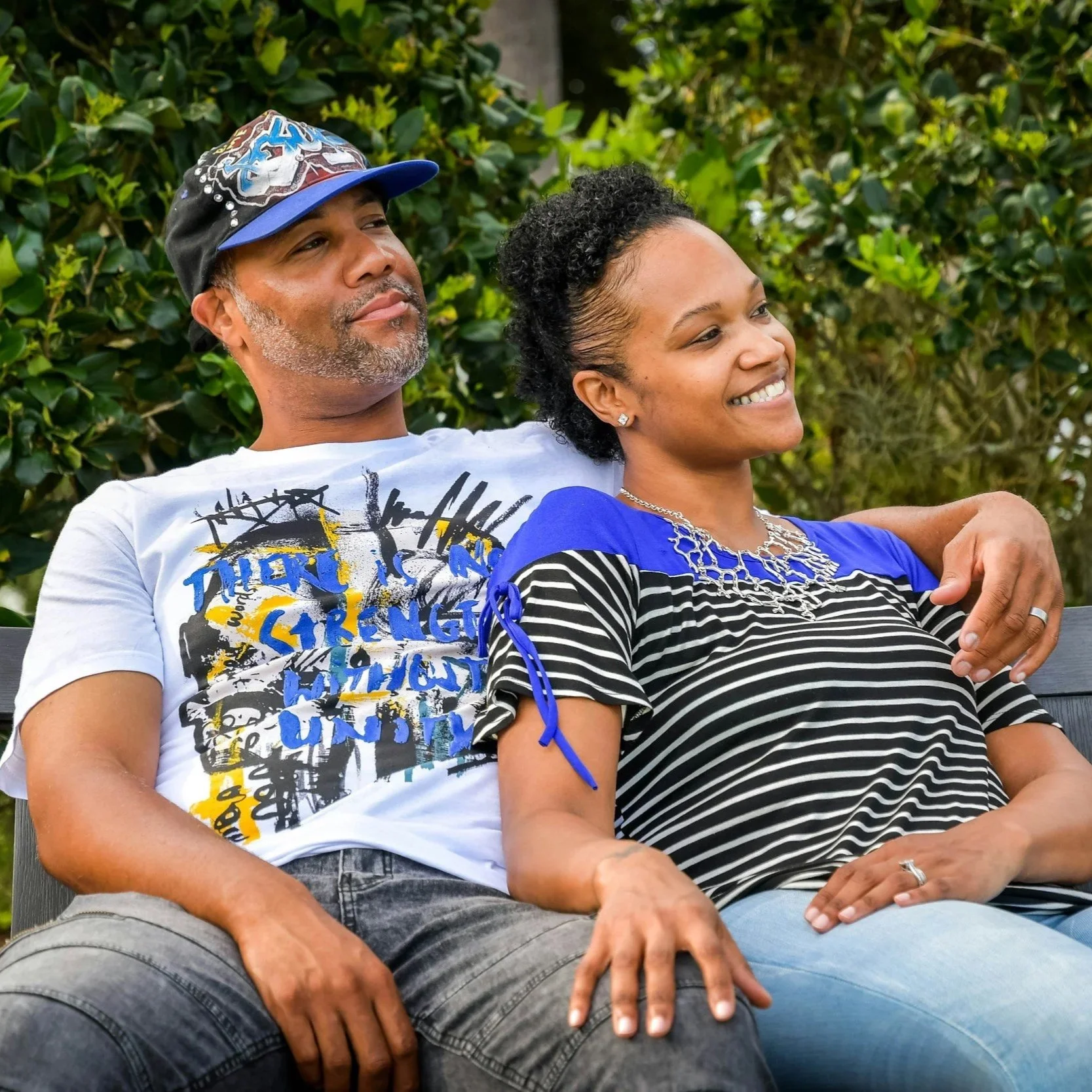 A man and woman sitting on a park bench, smiling, with green bushes in the background. The man is wearing a cap and graphic T-shirt, and the woman is wearing a striped shirt and jewelry.