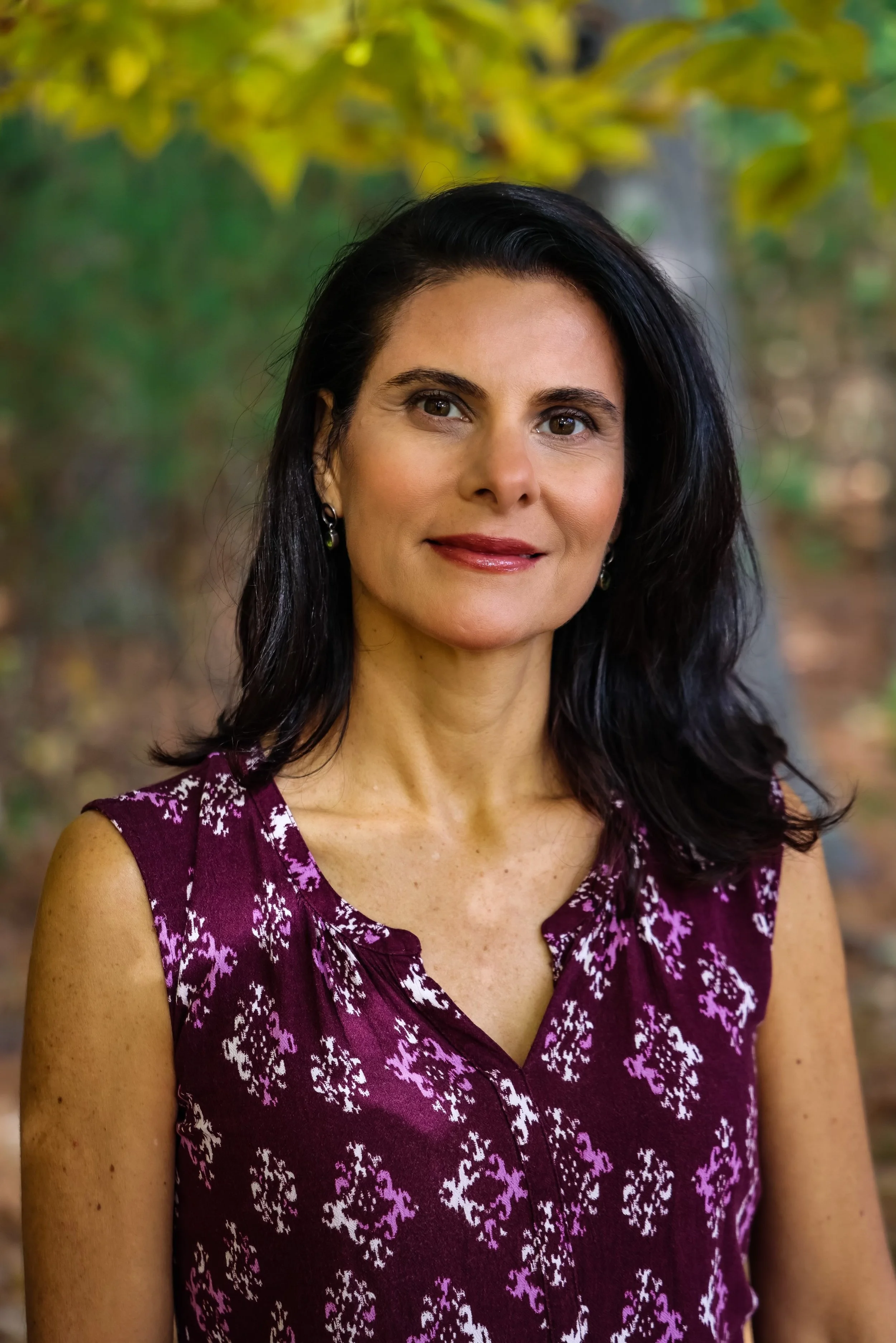 A woman with dark hair wearing a sleeveless purple top with white and purple floral patterns, standing outdoors with blurred trees and foliage in the background.