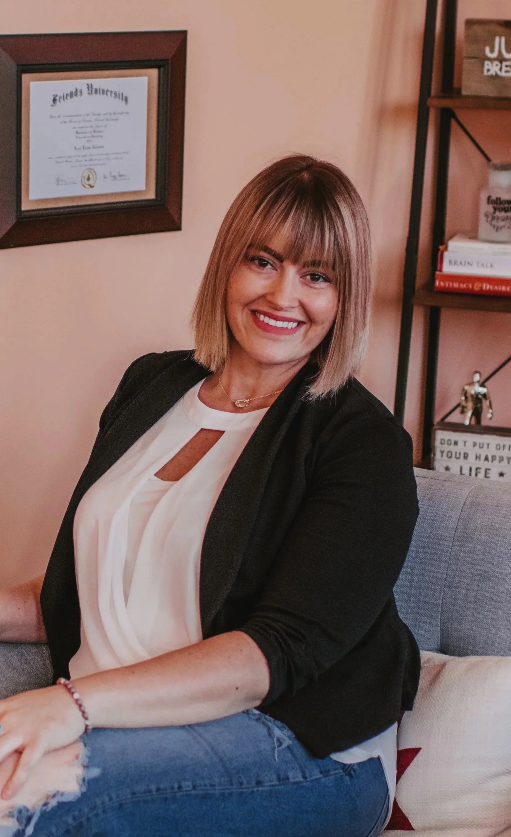 A woman sitting on a sofa, smiling at the camera, with a framed diploma on the wall behind her and a bookshelf with books and decor items.