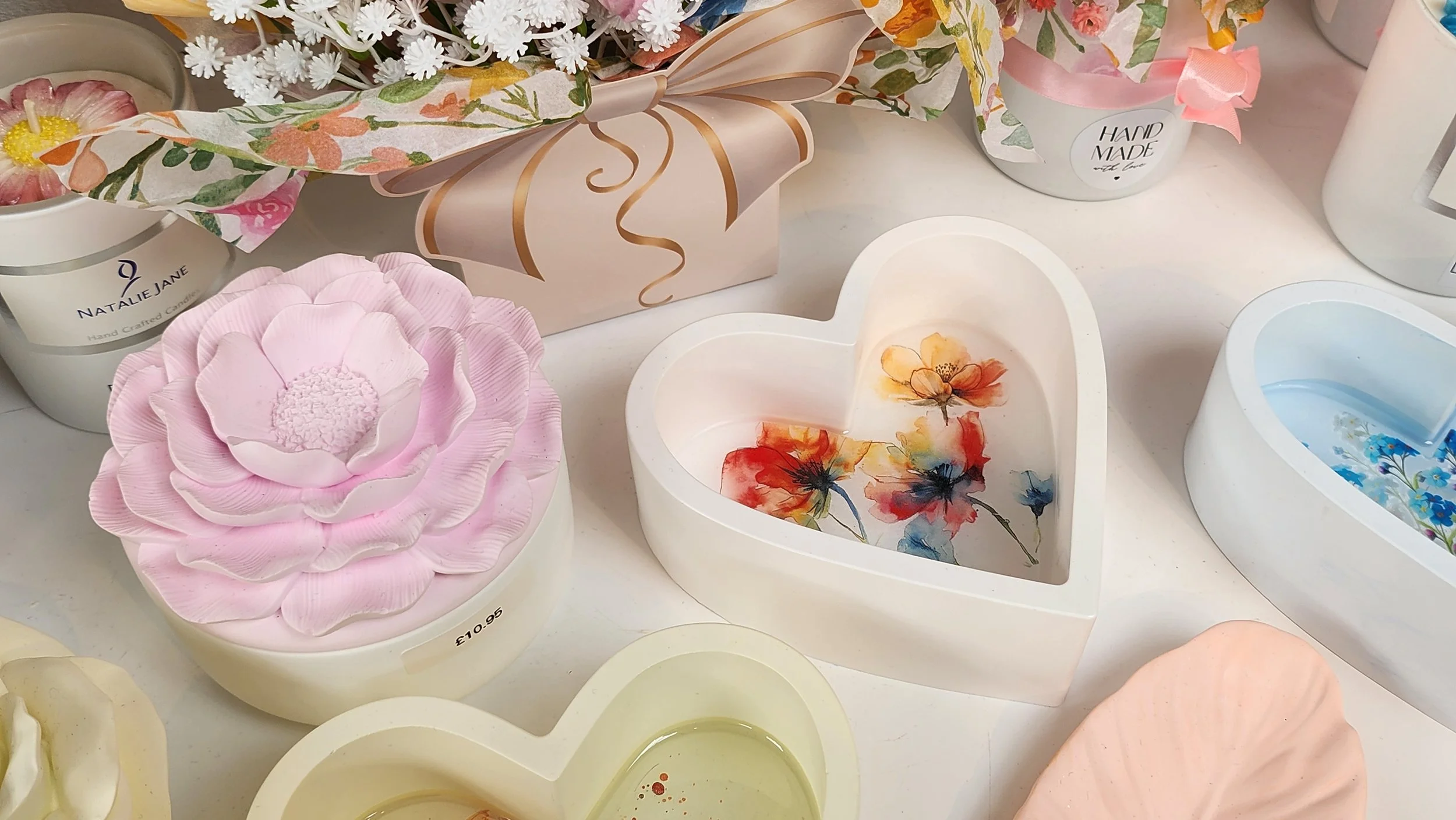 Various decorative gifts, including a pink flower-shaped eco-resin box, floral heart-shaped ornaments, and candles in containers with floral and handmade designs, arranged on a white surface.