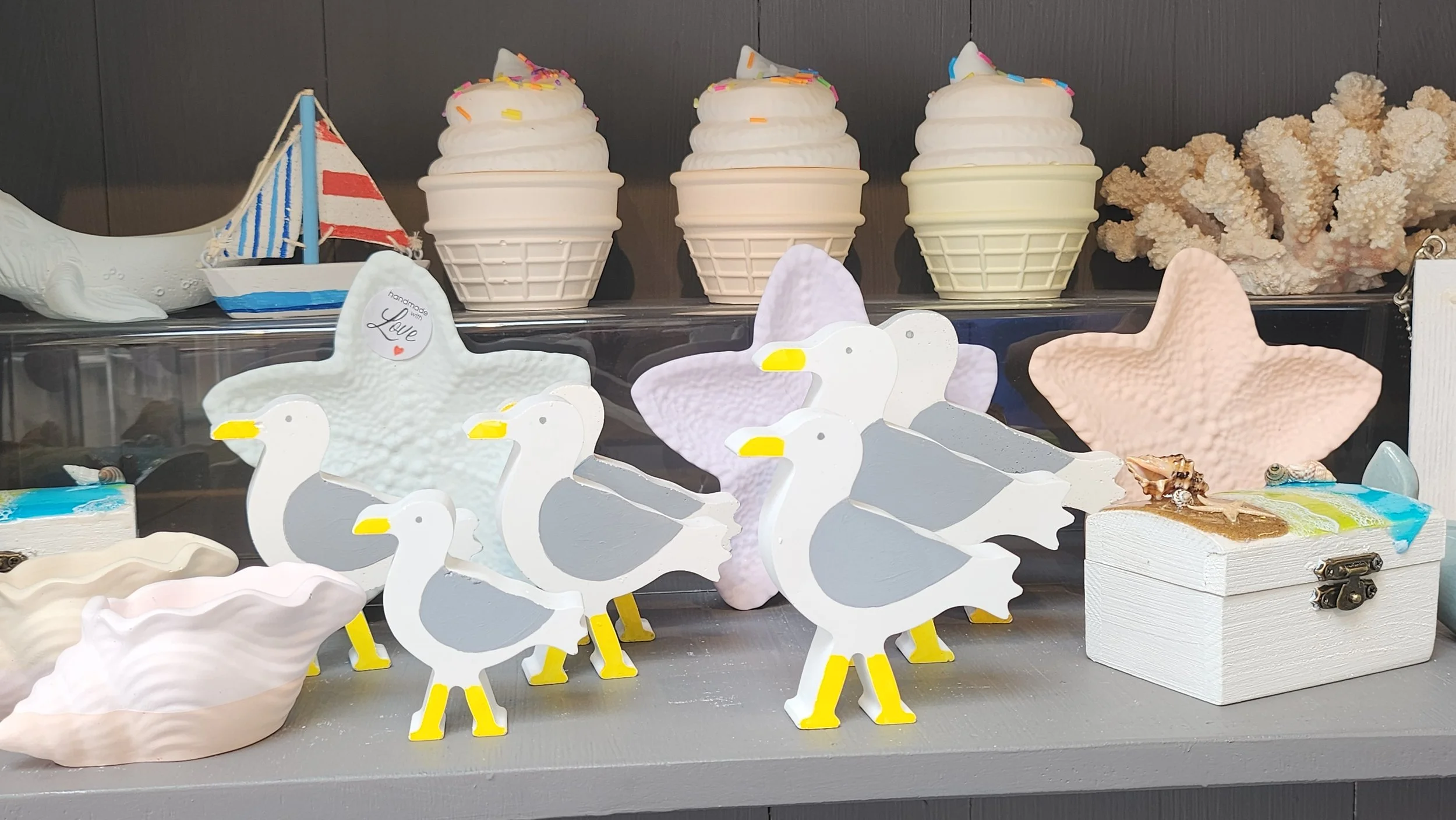 Decorative scene with wooden seagulls figurines, small toy sailboat, starfish and ice cream cone resin gifts on a shelf.