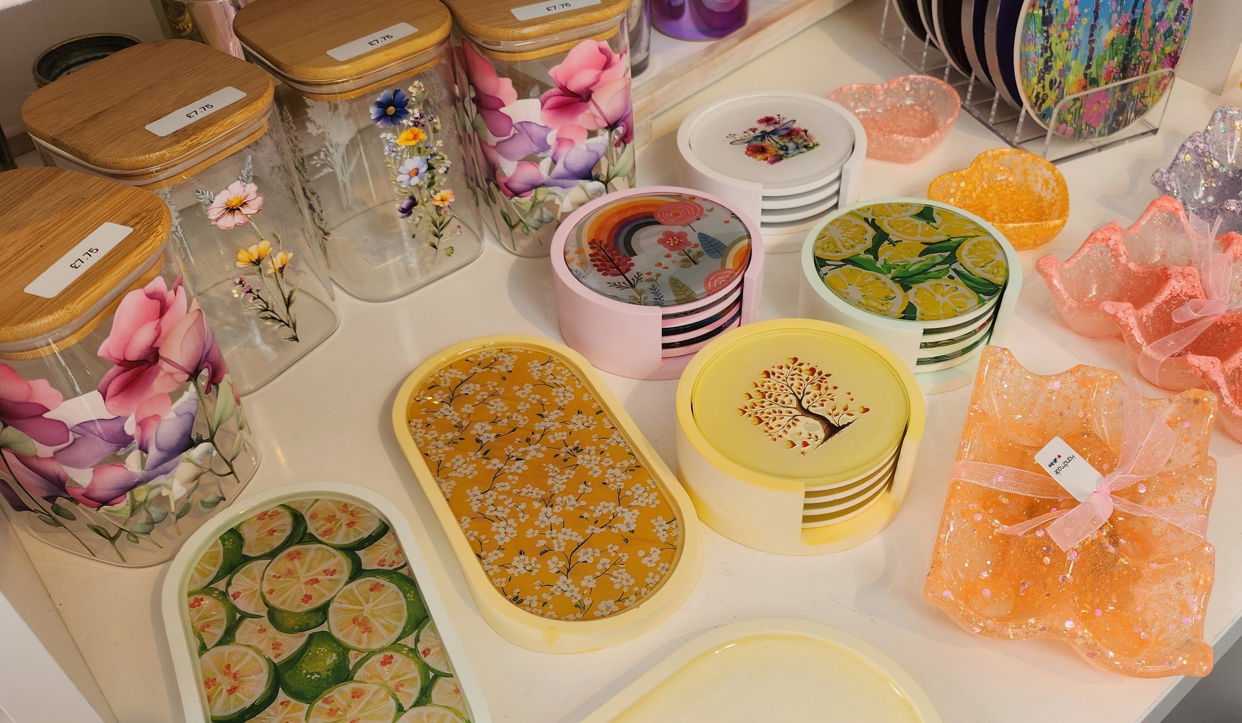 A display of colorful, decorative storage containers, bowls, resin coaster sets and small gift boxes with floral, lemon, rainbow, and tree designs, arranged on a table.