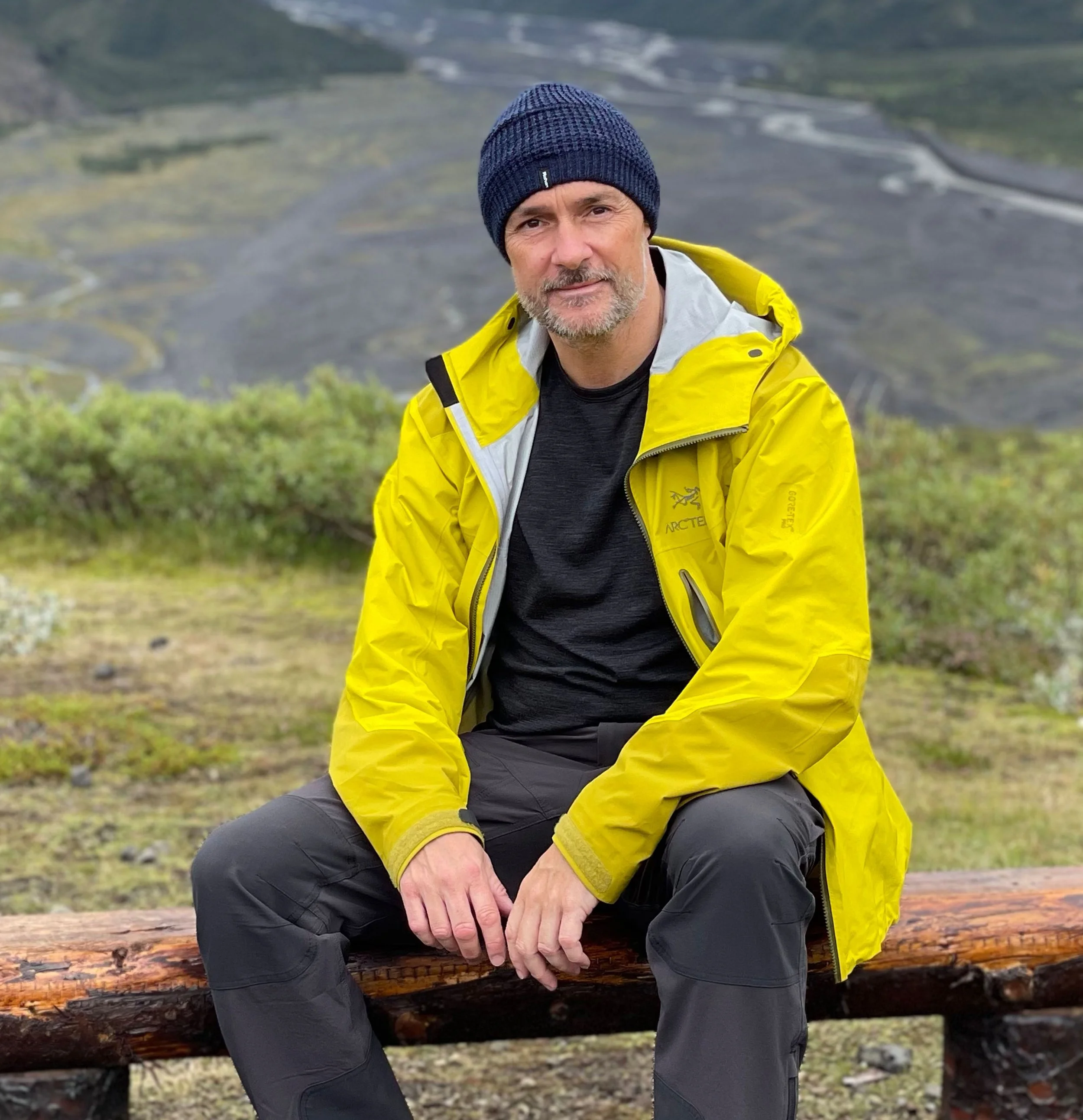 A man wearing a navy knit beanie, yellow outdoor jacket, black shirt, and black pants sitting on a wooden bench outdoors with a mountain and winding river in the background.