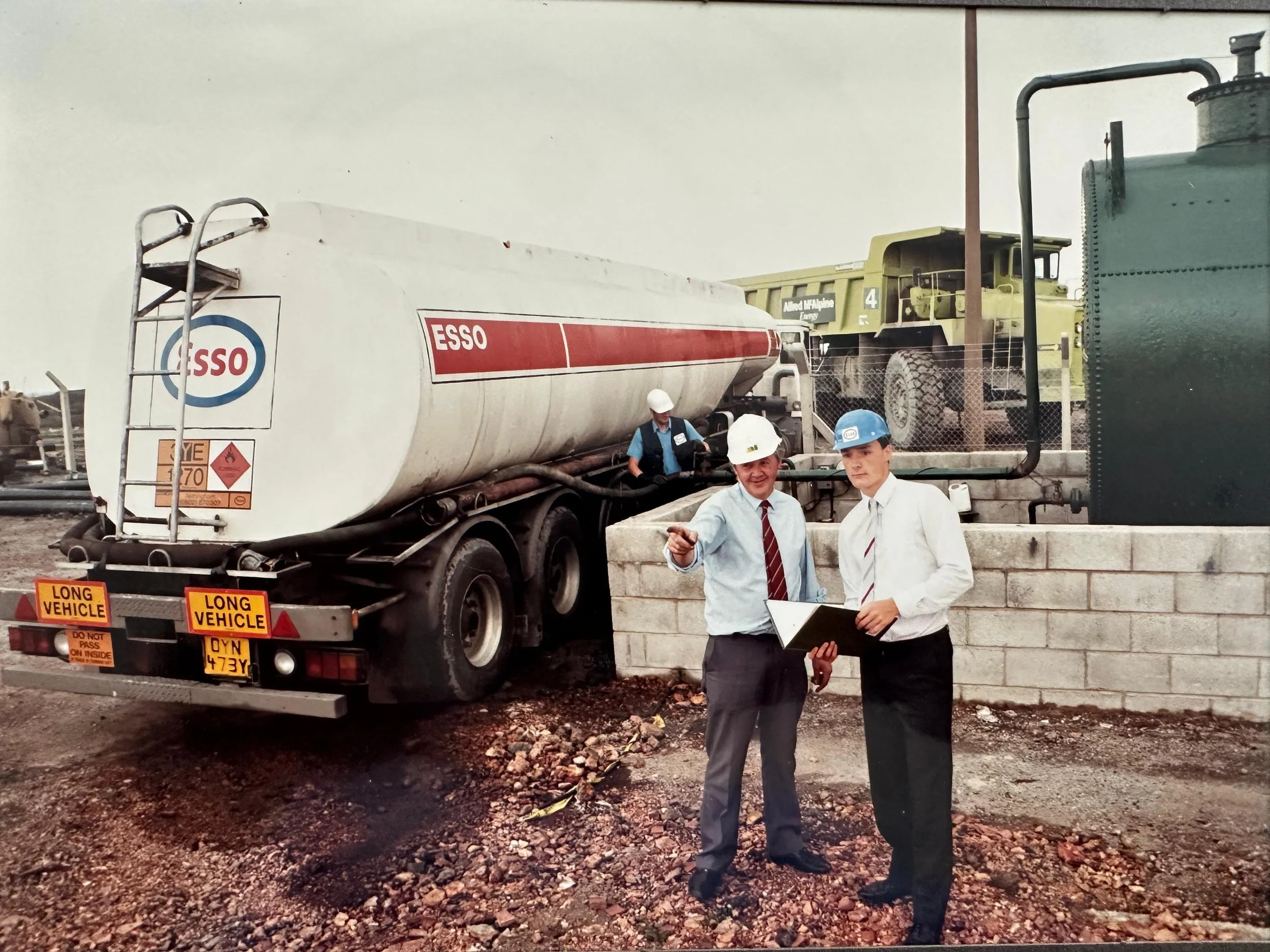 Two men in business attire and safety helmets stand near a concrete wall at an industrial site. One man is pointing while the other holds a clipboard. Behind them, a large tank truck branded 'ESSO' and a yellow heavy-duty truck are parked. The area has a gravel surface.
