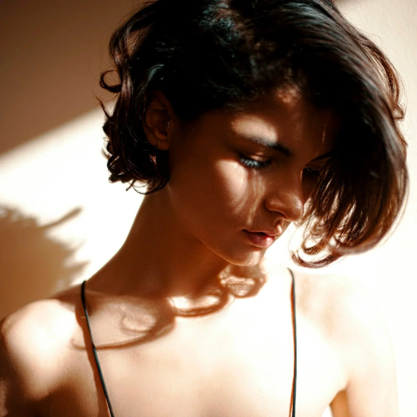 A young woman with dark hair and closed eyes looking downward, wearing a thin strap top, with soft lighting and shadow patterns on her face and shoulder.